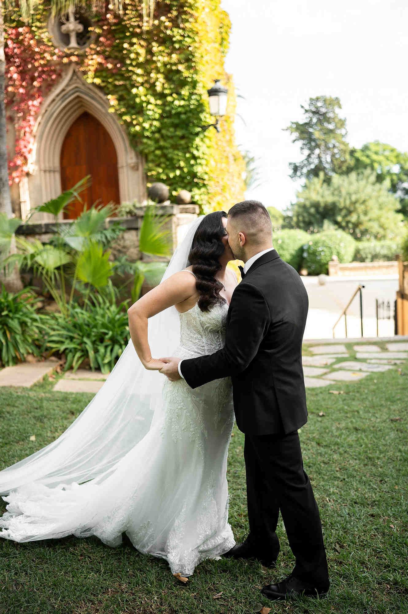 Romantic first kiss of the newlyweds in the sun-drenched Mediterranean gardens of the Casa Santonja estate.