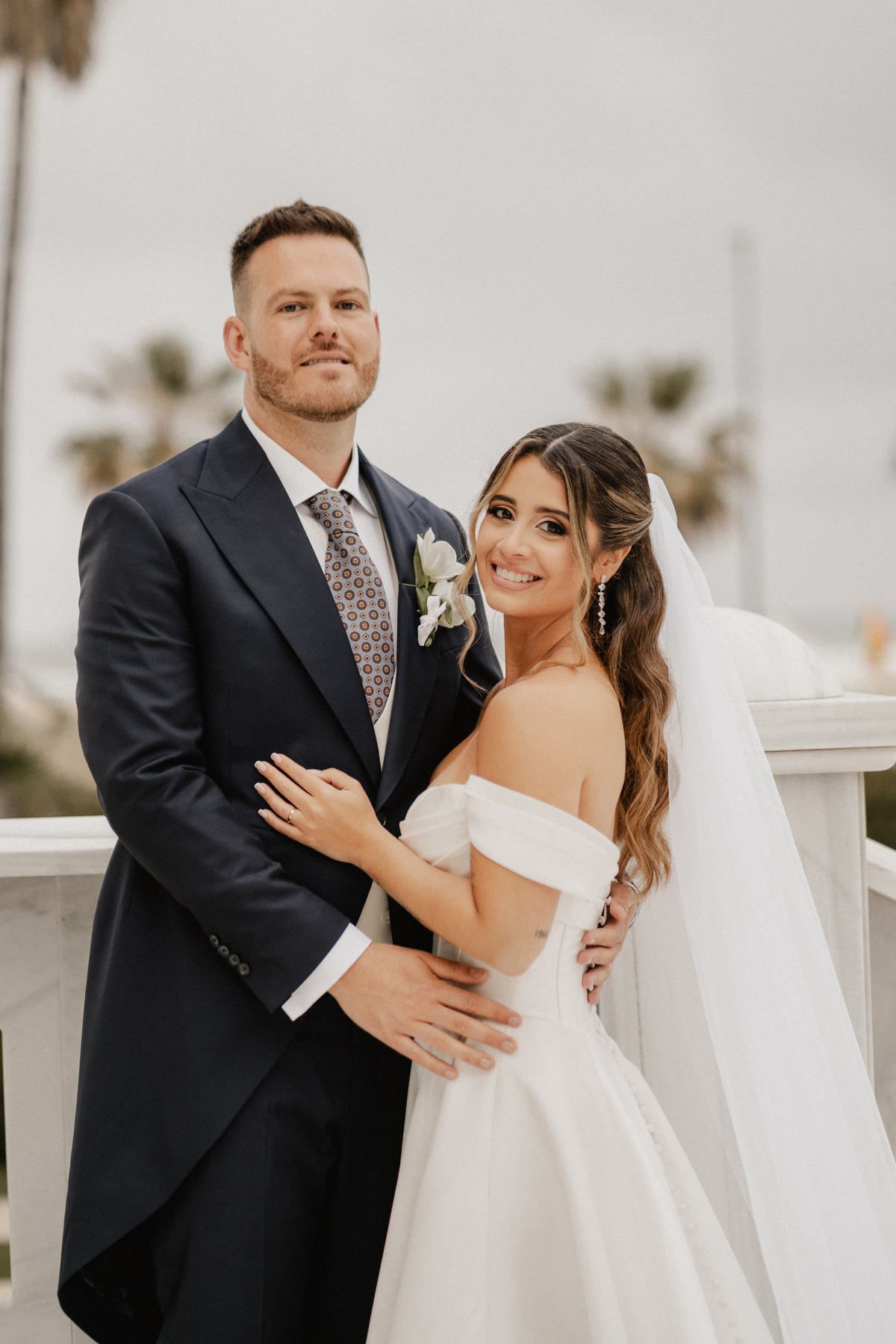 Bride and groom posing by the white marble balustrades overlooking the palms at Las Arenas; her half-up style looks flawless.