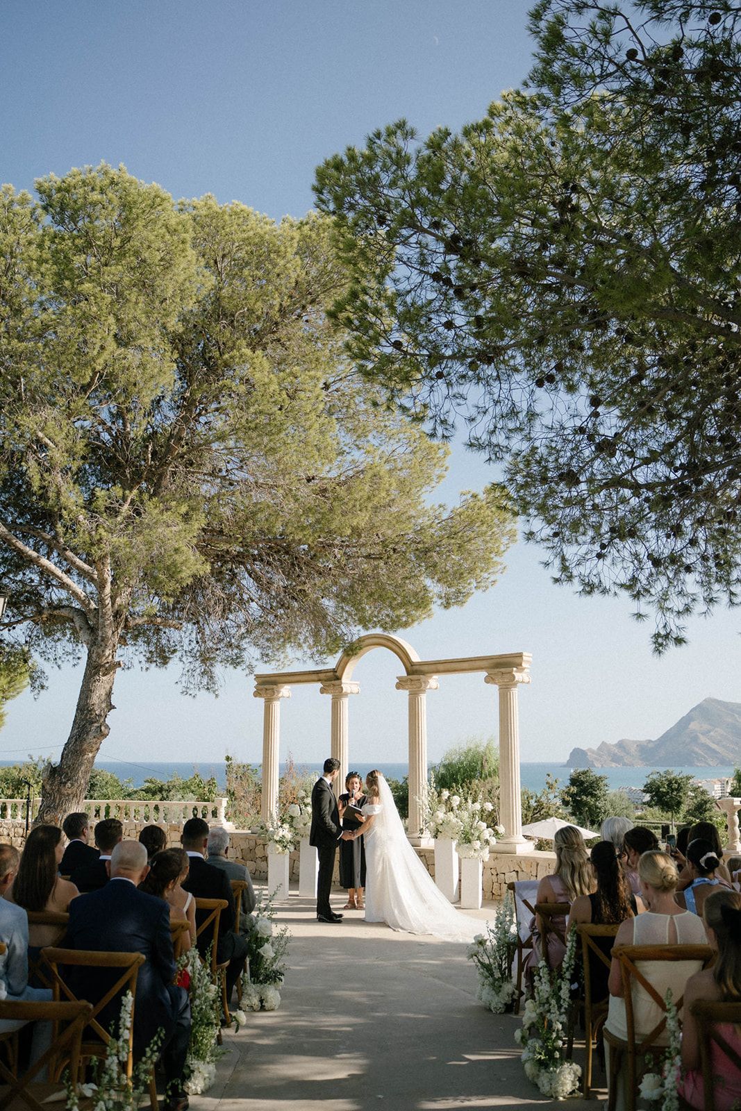 Wide angle shot of the bride and groom at the wedding ceremony surrounded by guests at the scenic Finca Marqués de Montemolar in Altea.