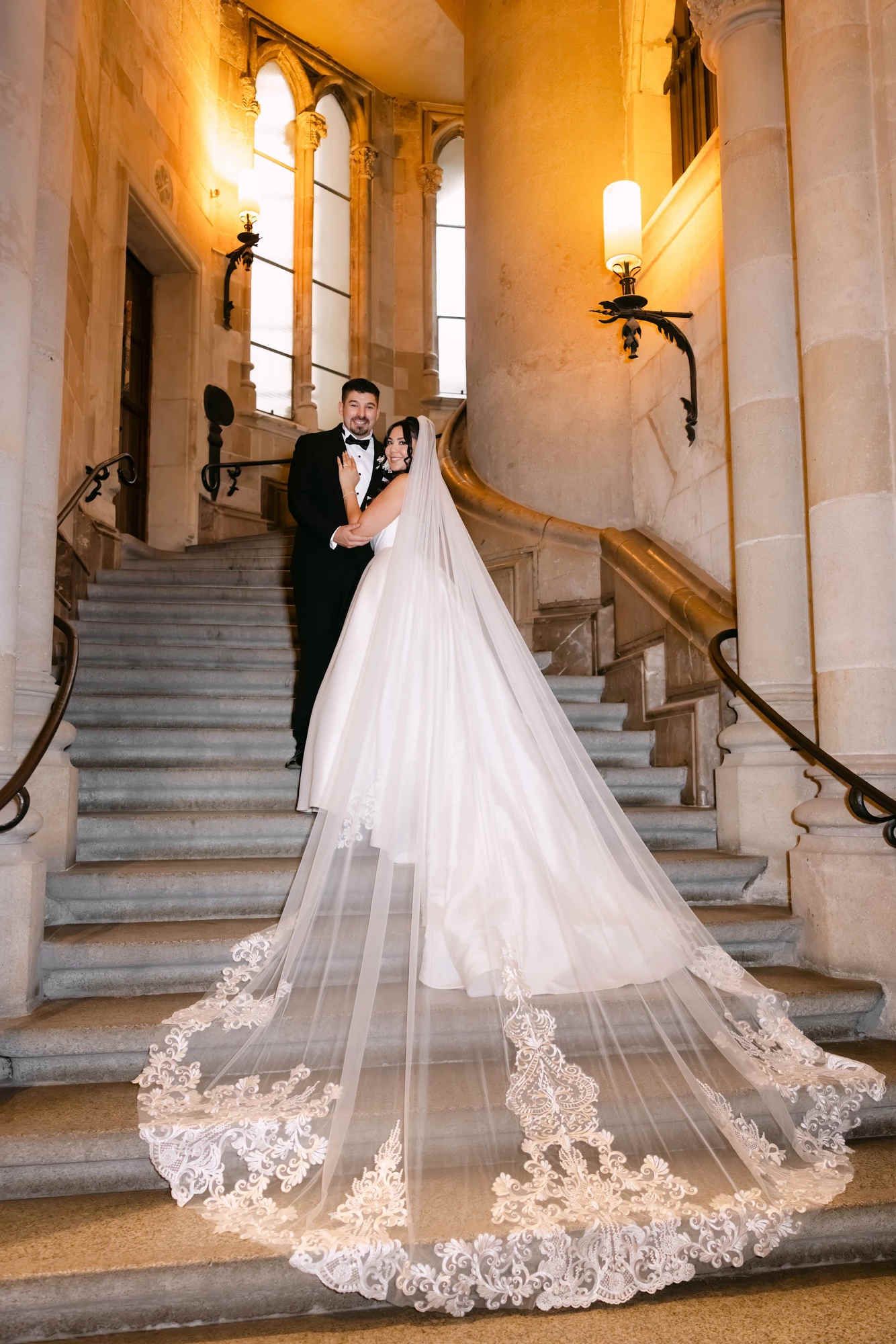 Full-length portrait of the newlyweds inside Sagrada Família, showing the long bridal train draped over the stairs.
