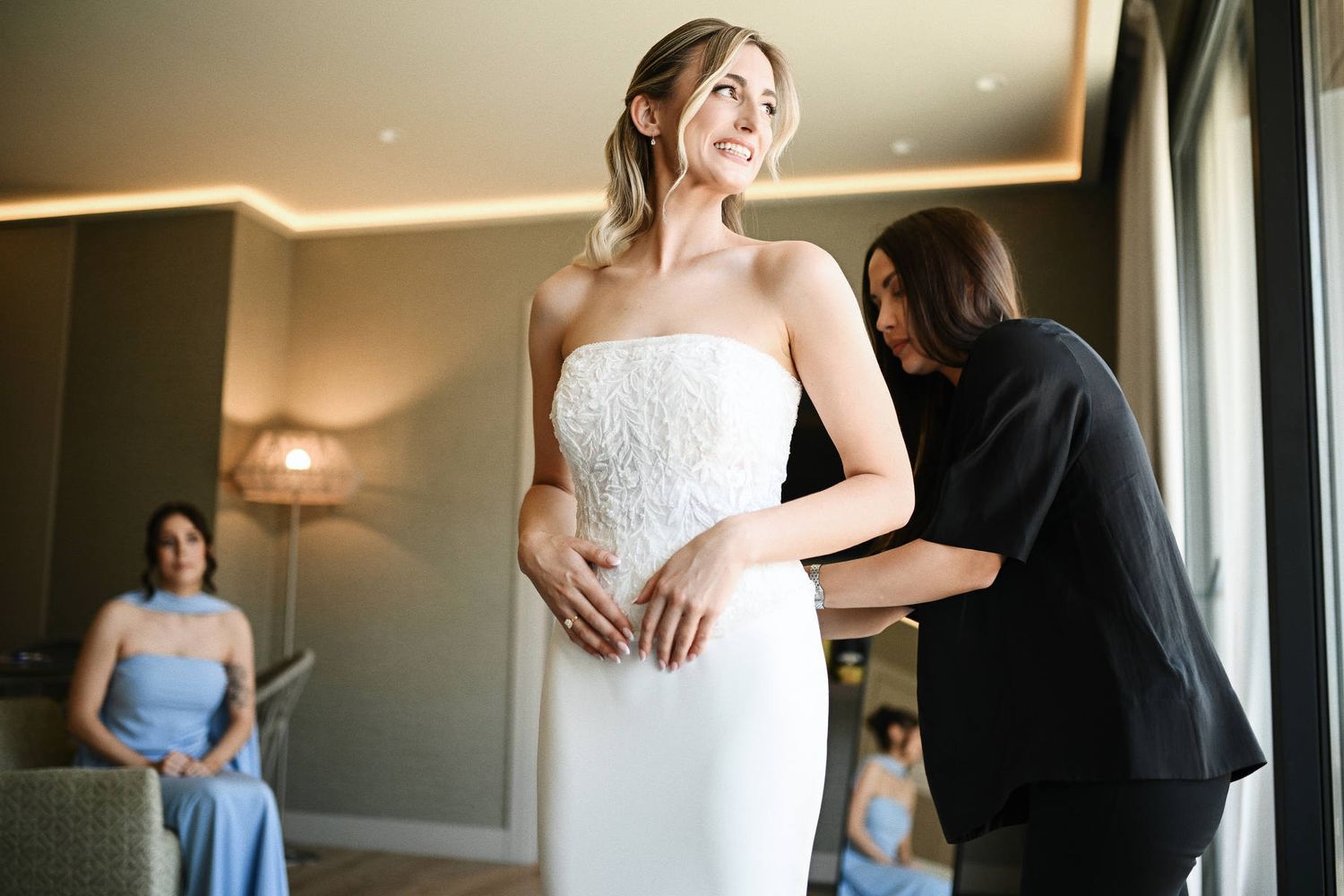 Professional beauty stylist helping the bride lace up her wedding gown, finishing the luxury bridal look inside the Villa Delia manor.