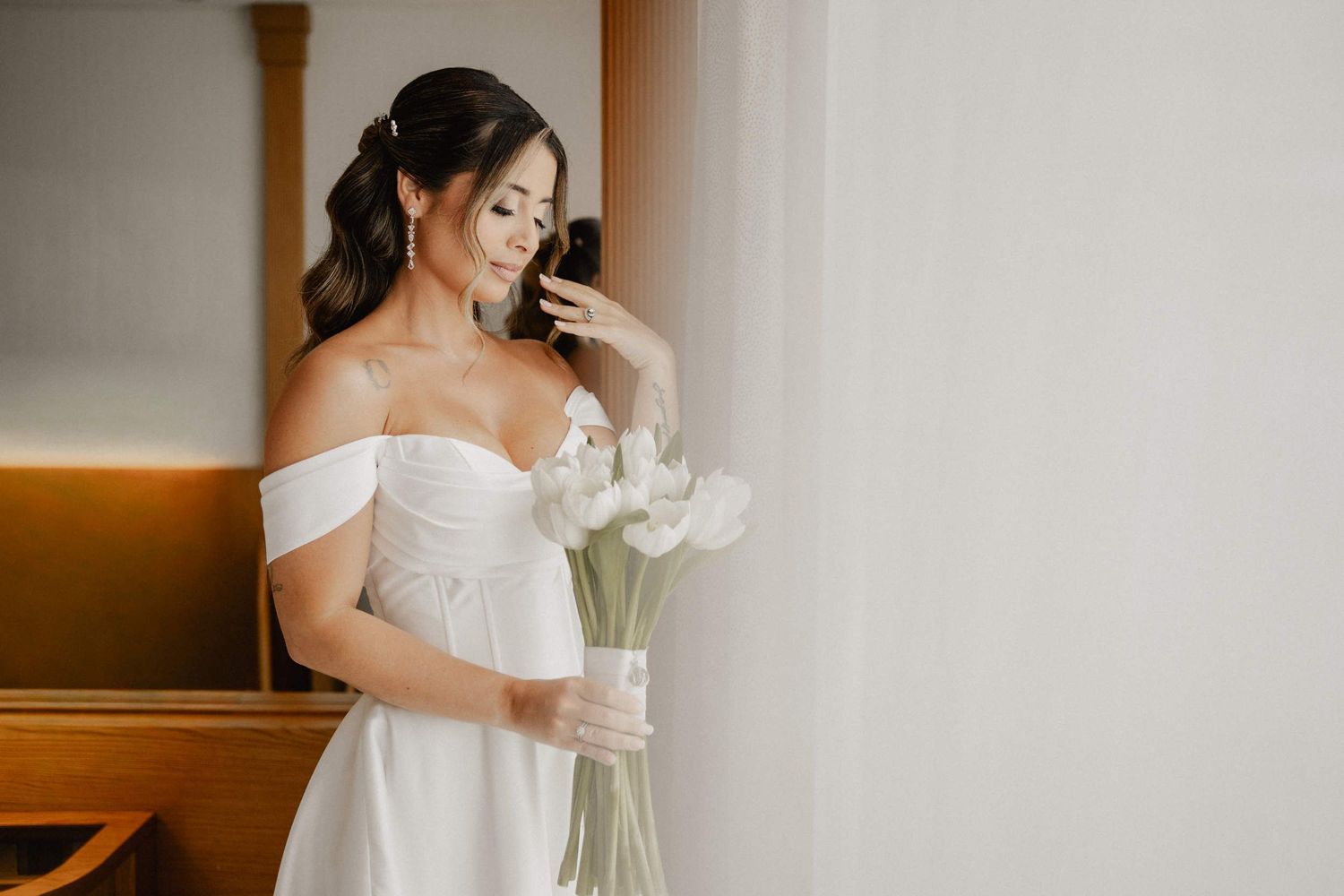 Bride in a light-filled suite at Las Arenas with polished half-up waves, defined eyes, and a nude satin lip holding white tulips.