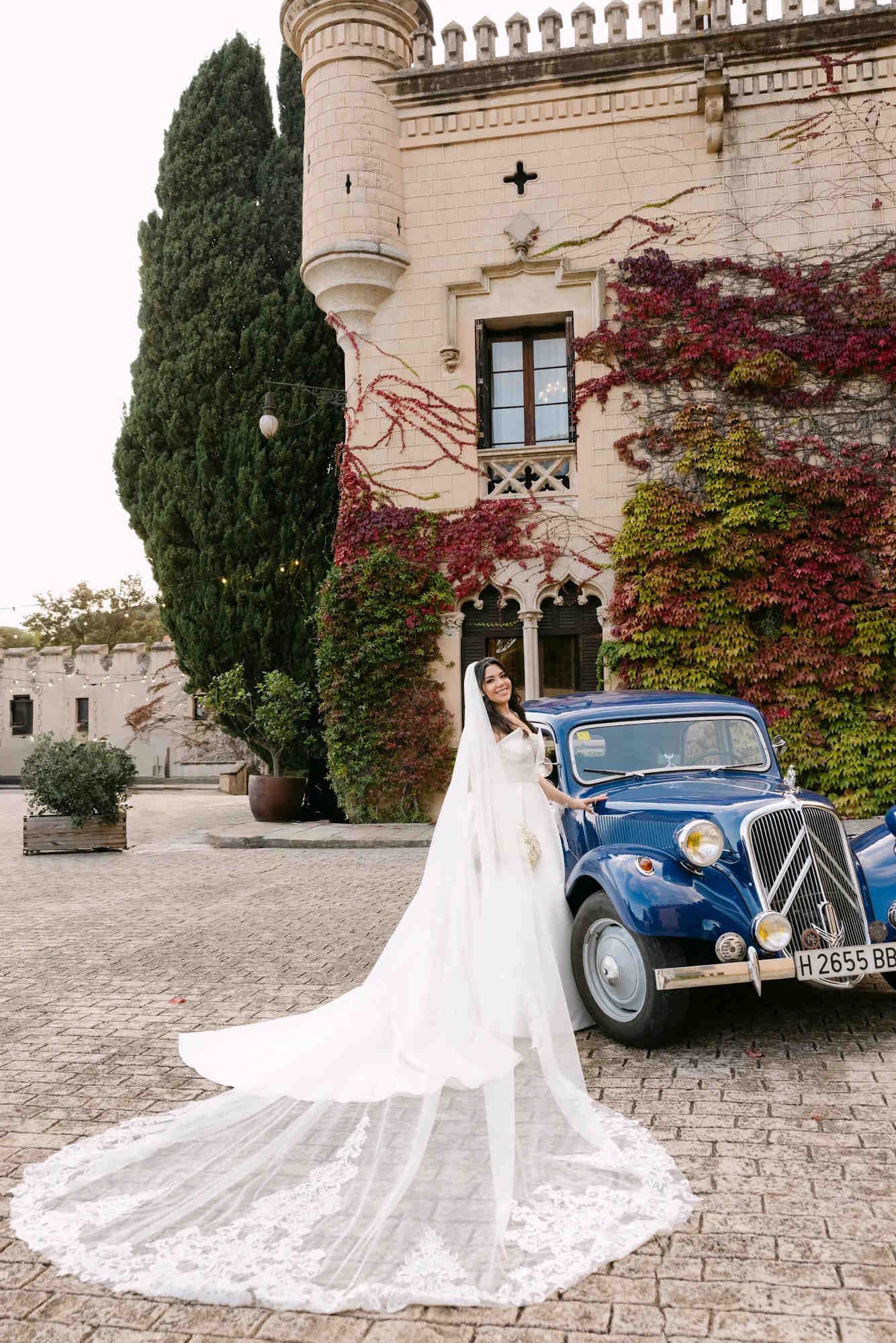 The bride posing with a classic blue vintage car and a long dress train against the majestic backdrop of Castell Jalpí