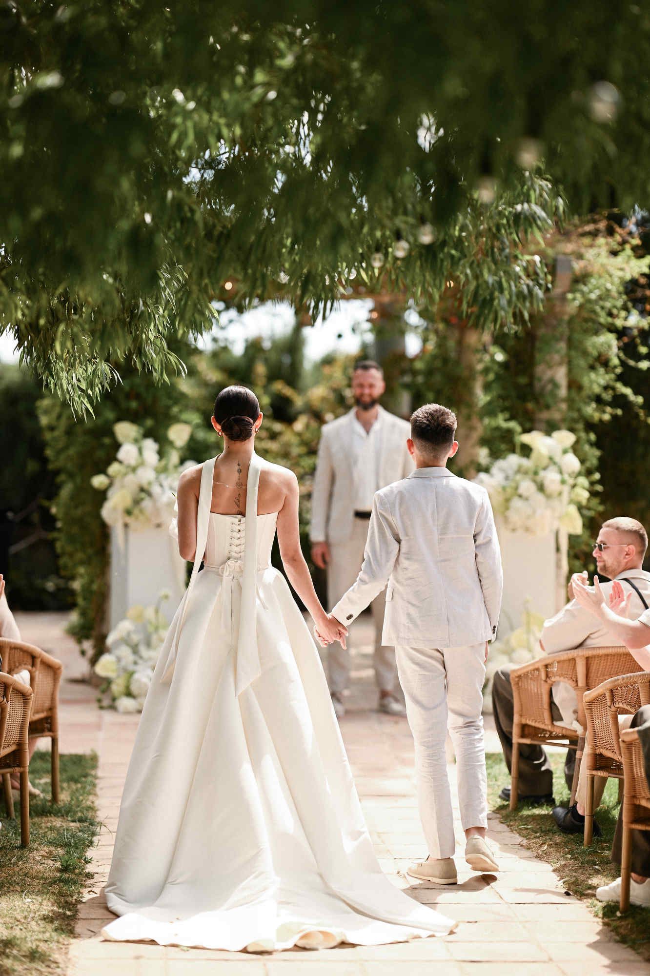 Back view of a son walking the bride to the altar, showing her bridal gown, dress train, and sleek bun hairstyle at Villa Paula.