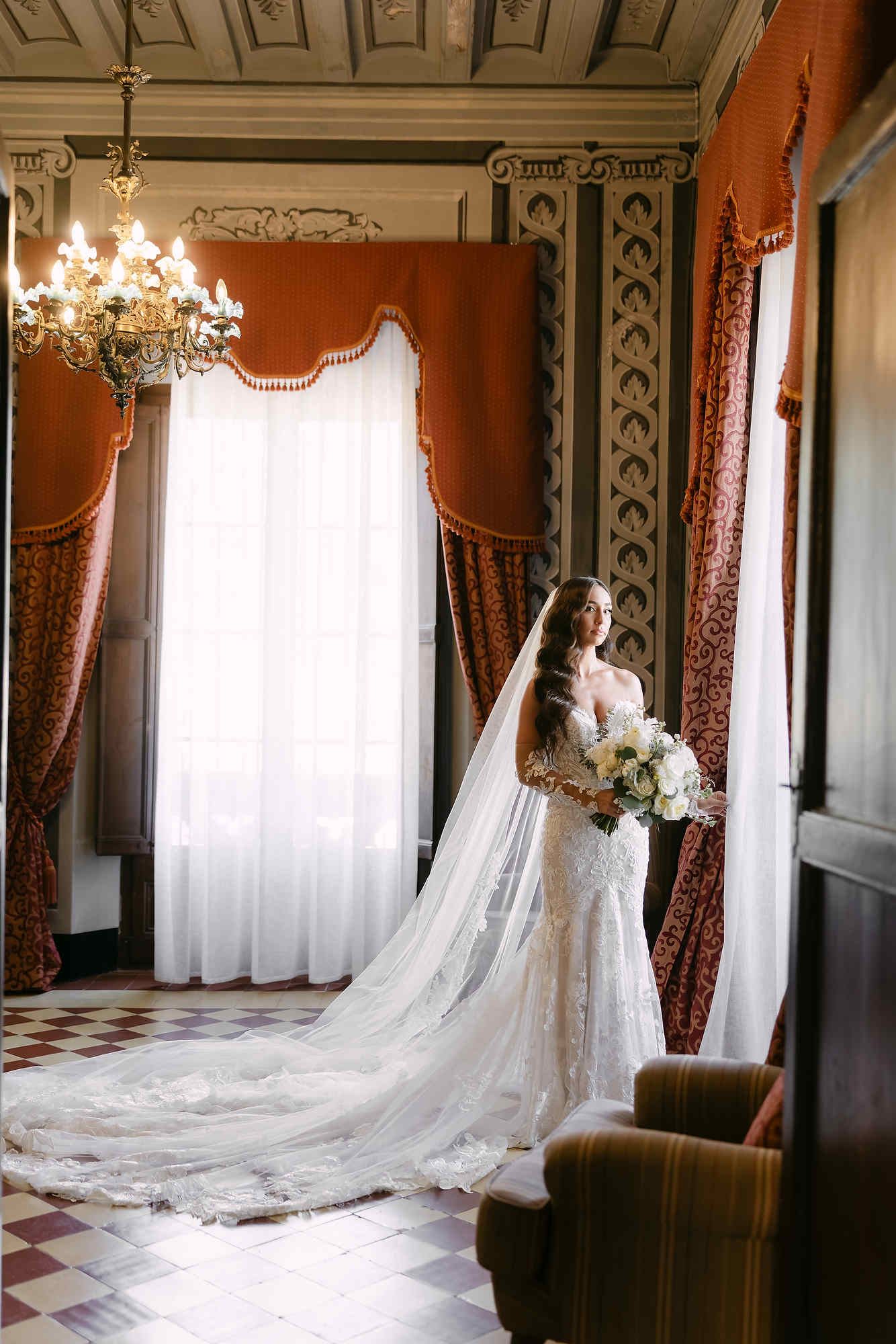 Wide shot of the bride with a long dress train looking out the grand window of Castell Jalpí, showcasing her elegant silhouette