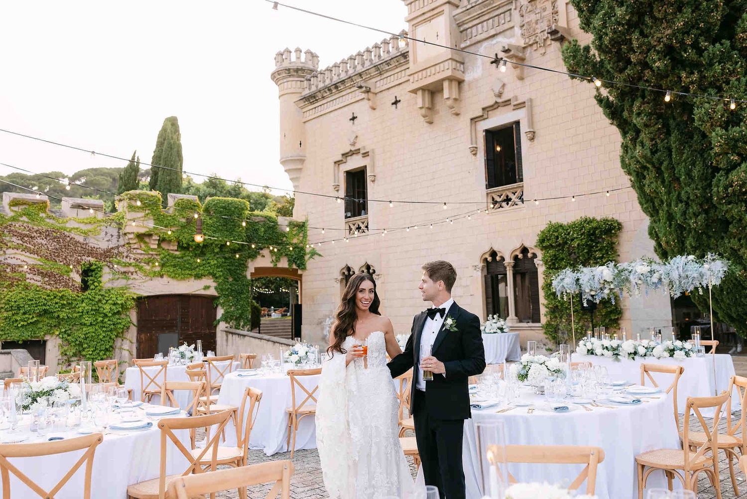 The bride and groom posing at Castell Jalpí with the luxury outdoor banquet tables and the historic castle facade in the background