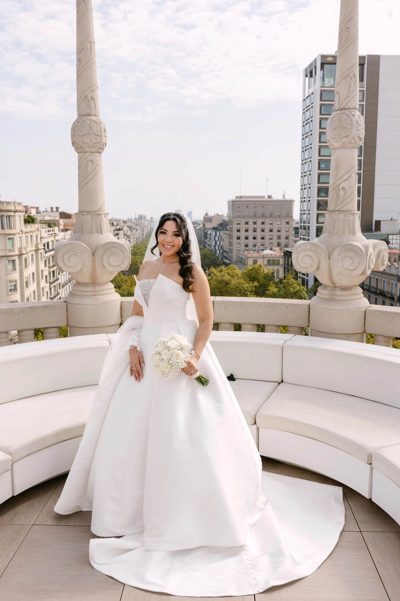 Full-length bridal portrait on a Barcelona rooftop featuring the finished hair and makeup look with a panoramic city view.