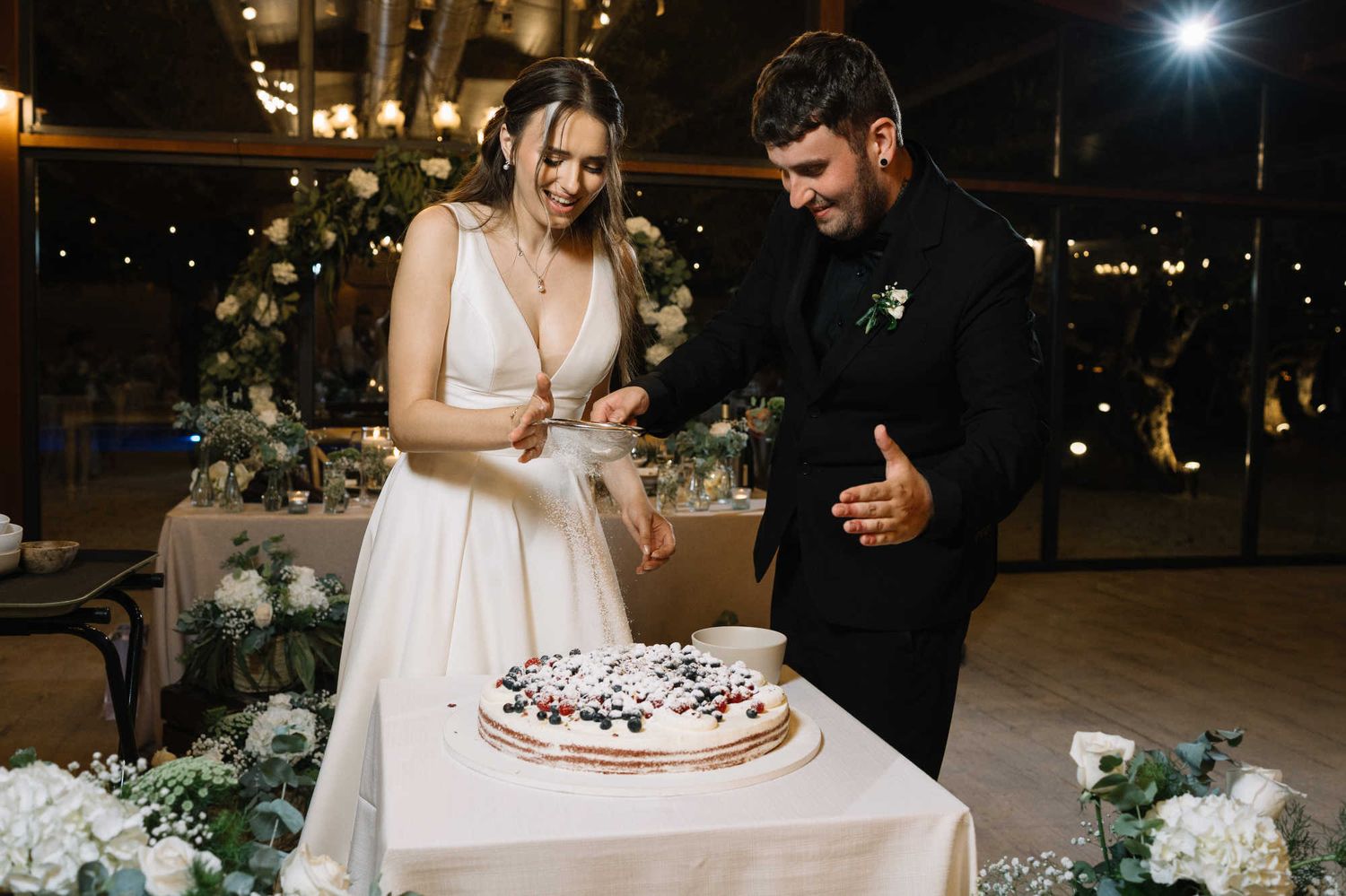 Bride and groom cutting the wedding cake under string lights during an evening reception at the rustic Alqueria del Xúquer estate in Valenci