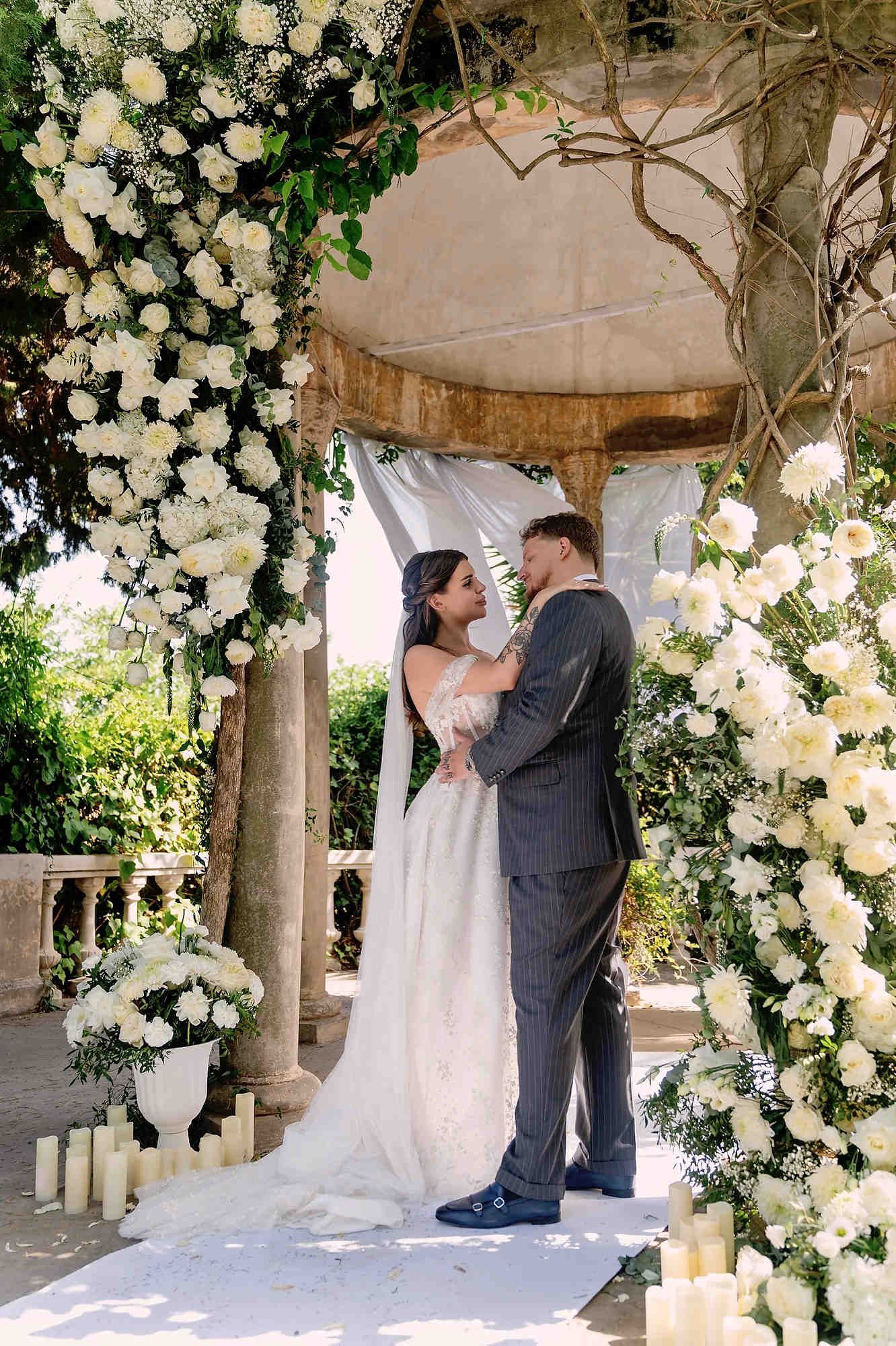 Groom and bride embracing under a luxury floral wedding arch during their ceremony at Casa Santonja.