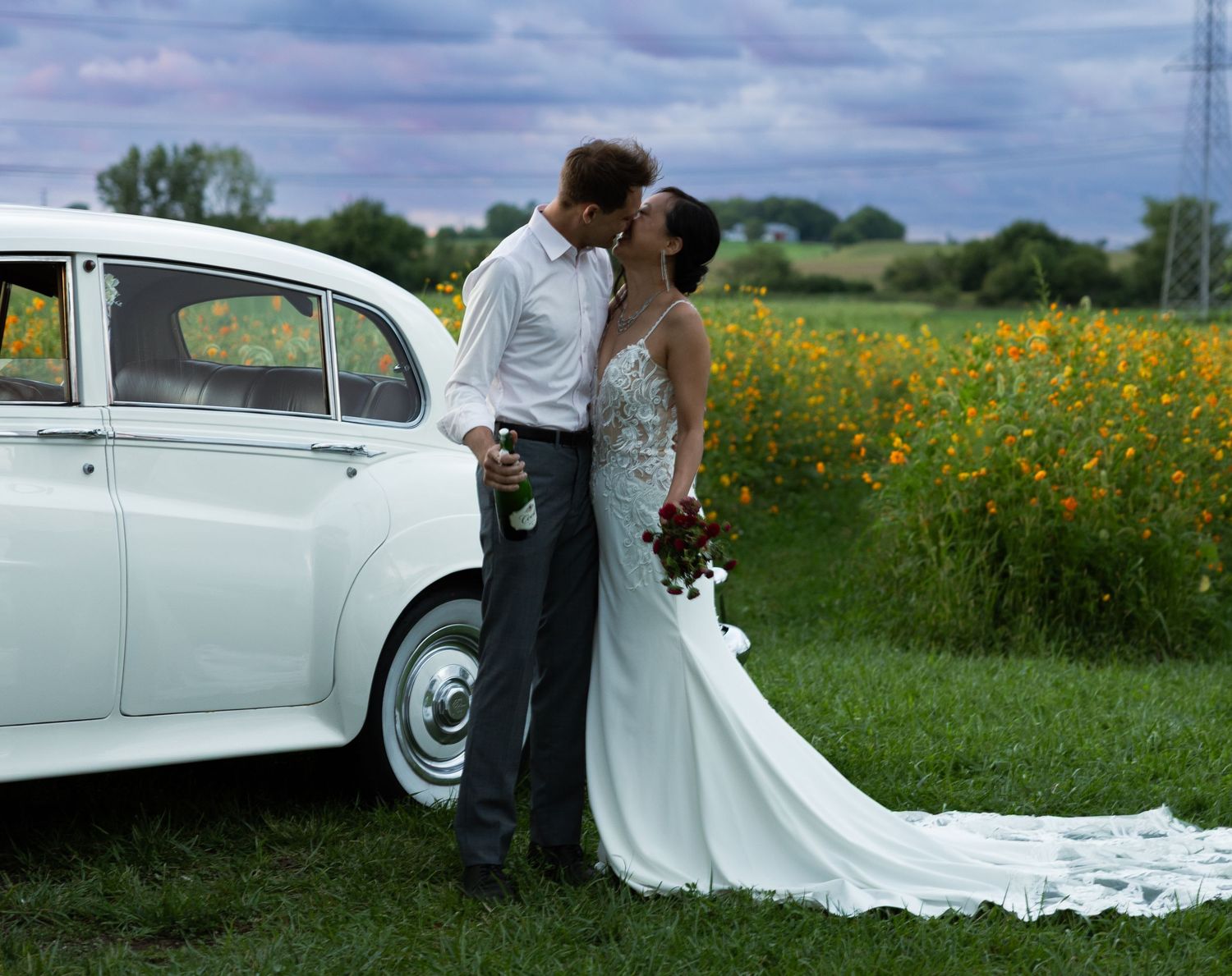 Newlyweds share a kiss beside classic white car in field of wildflowers.