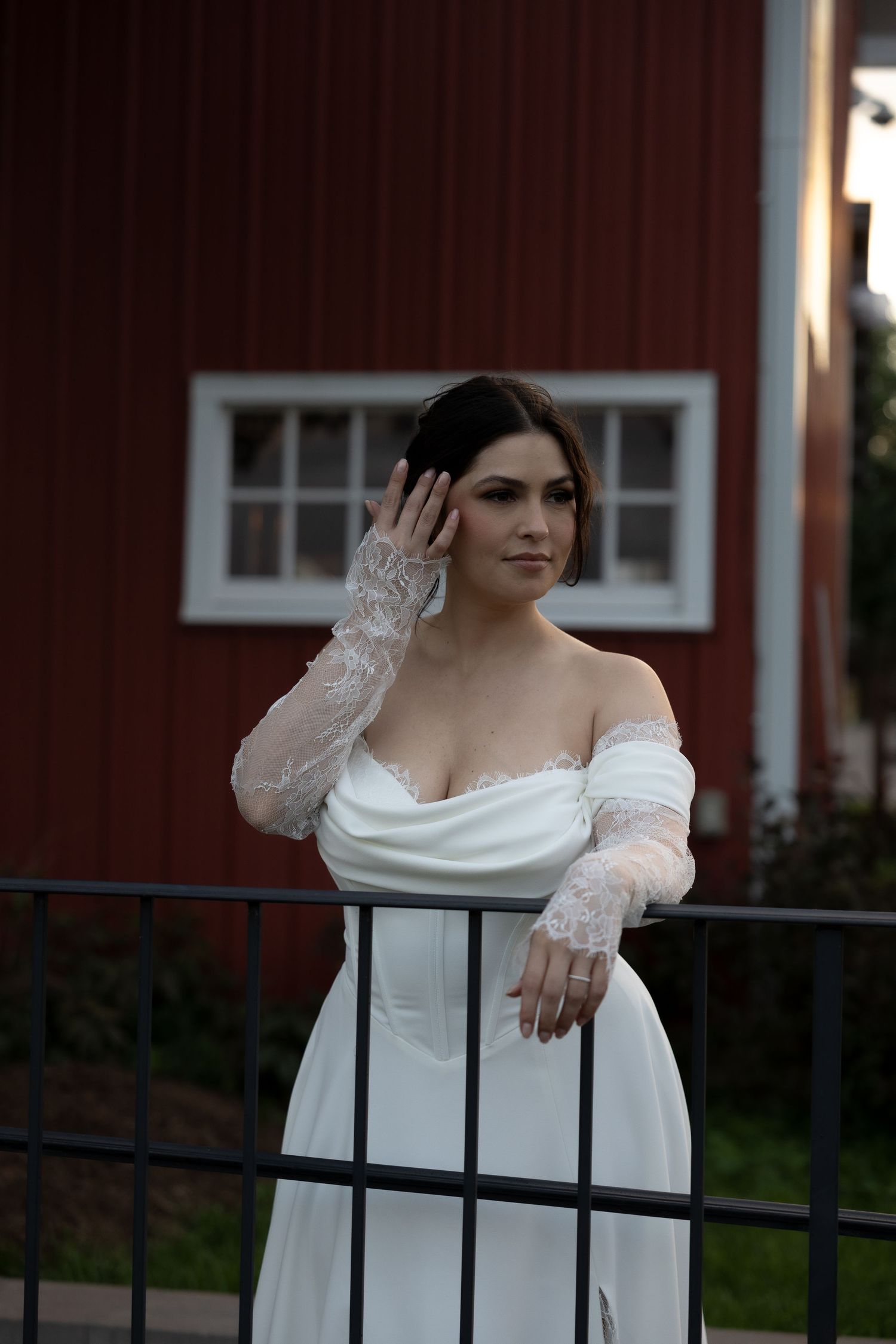 Person in white off-shoulder dress leans against dark railing in front of red building.