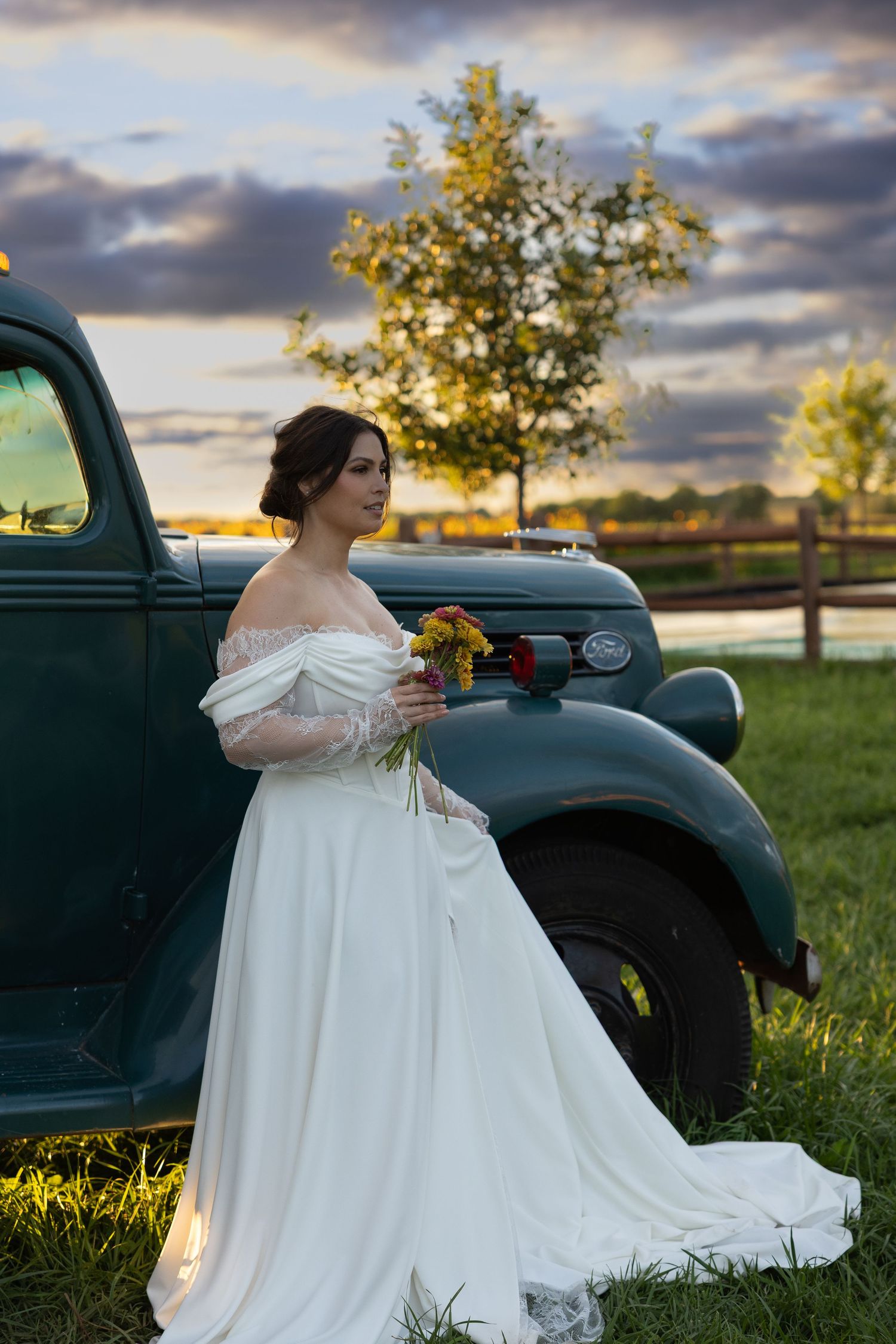 Bride in off-shoulder gown poses beside vintage truck during golden hour.
