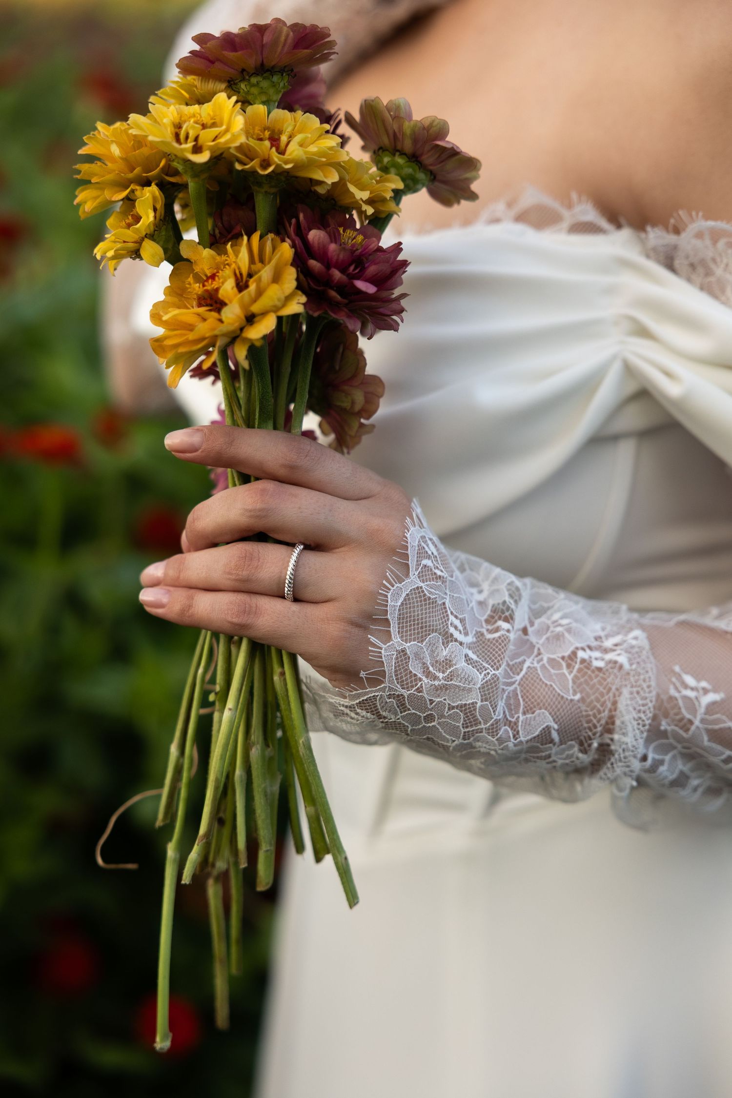 Close-up of bridal bouquet with yellow and burgundy flowers held by lace-sleeved hands with wedding ring.