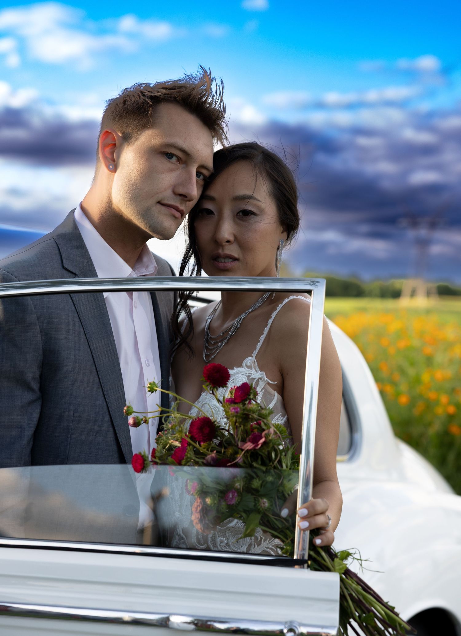 A romantic wedding portrait through a car window with red roses and yellow wildflowers in background.