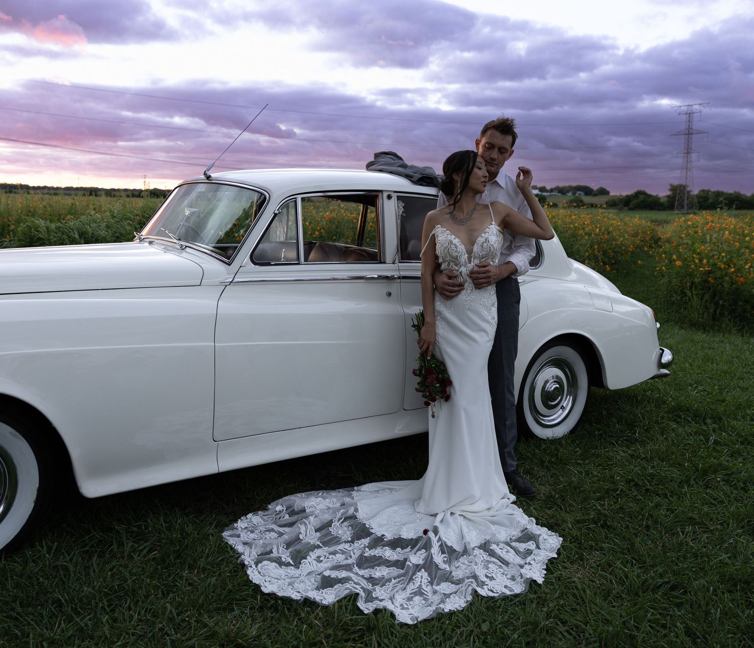 Elegant white vintage car against dramatic sky with wedding dress train on grass.