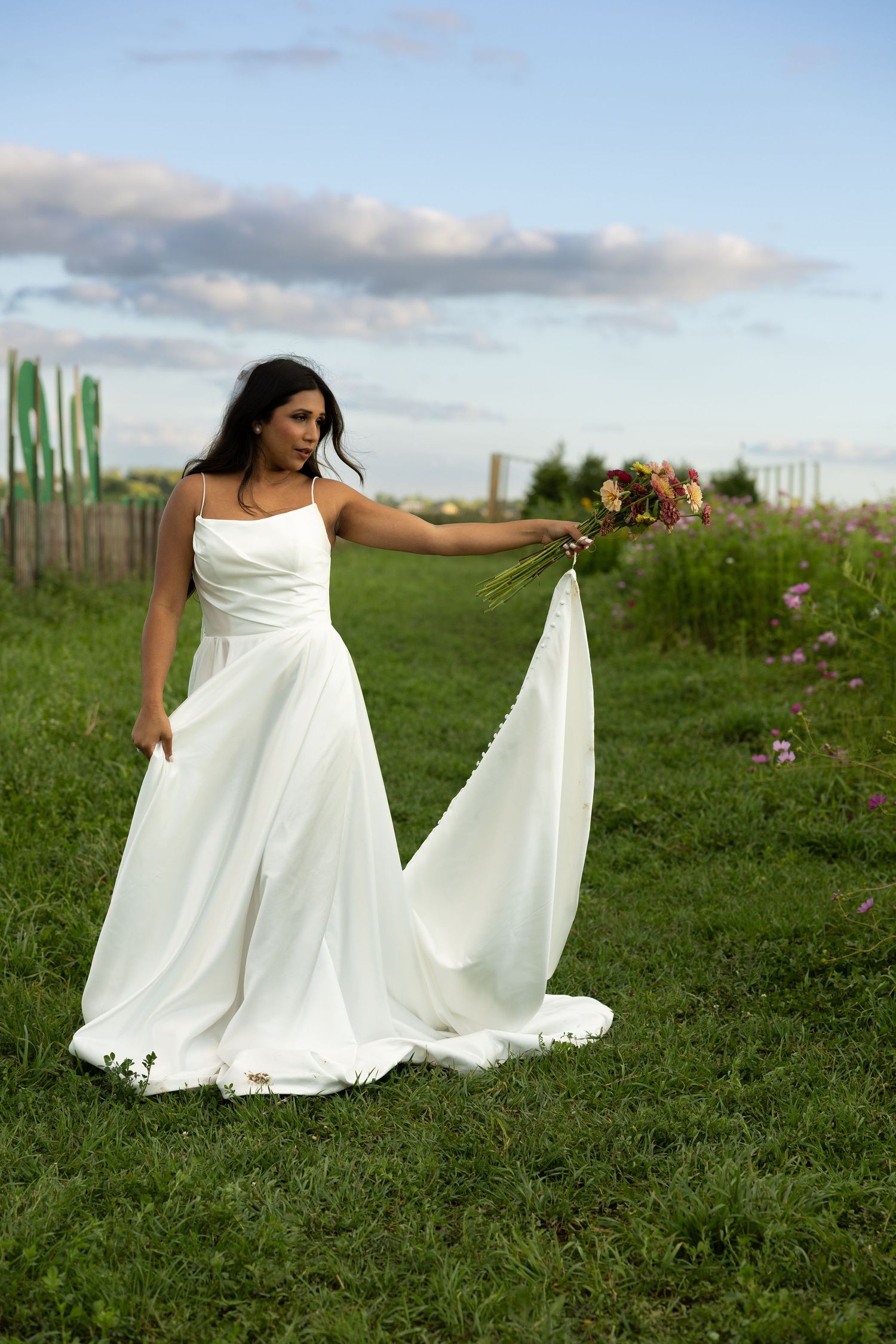 A bride in a flowing white wedding dress stands in a grassy field holding a bouquet against a cloudy sky.