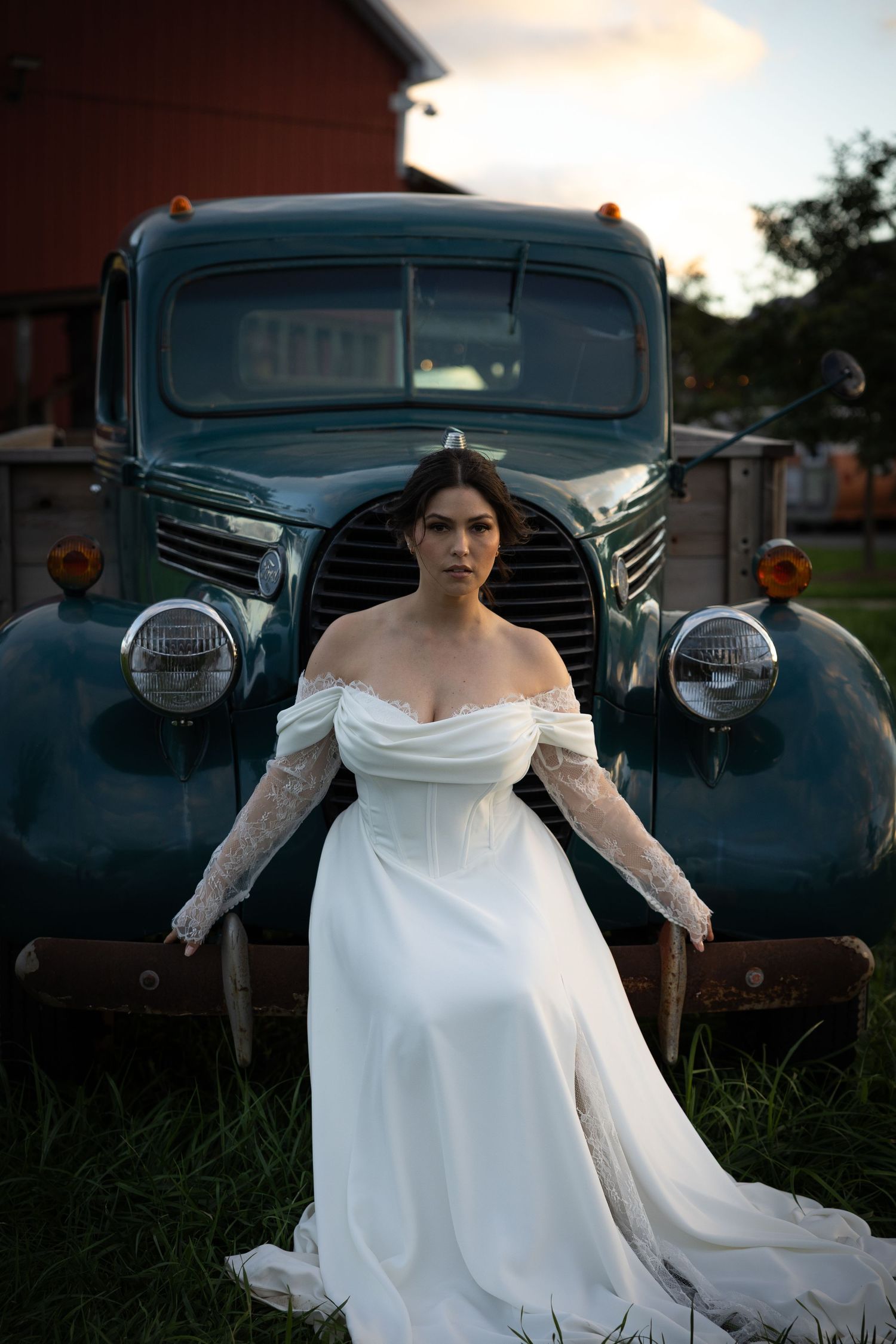 A bride in a white off-shoulder gown poses dramatically against a vintage black car at dusk.
