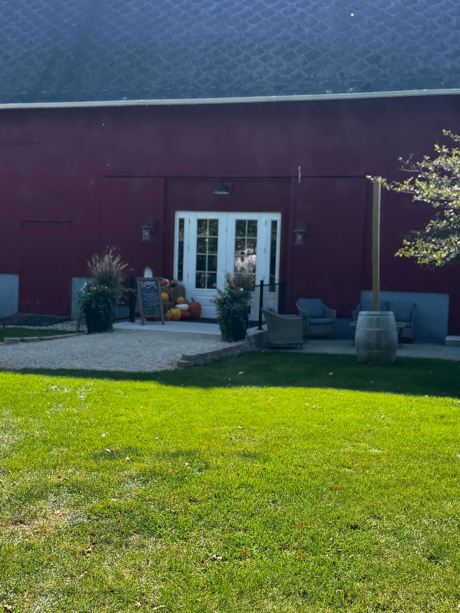 Red barn venue with white French doors, fall pumpkin decor, wooden barrel, and lush green lawn on a sunny day.