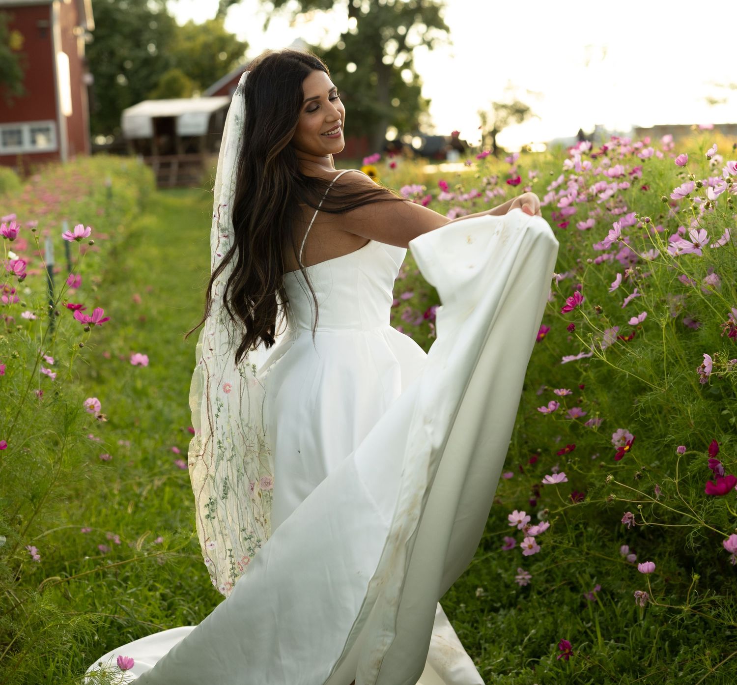 A bride in a flowing white dress twirls gracefully in a garden filled with pink cosmos flowers on a summer day.