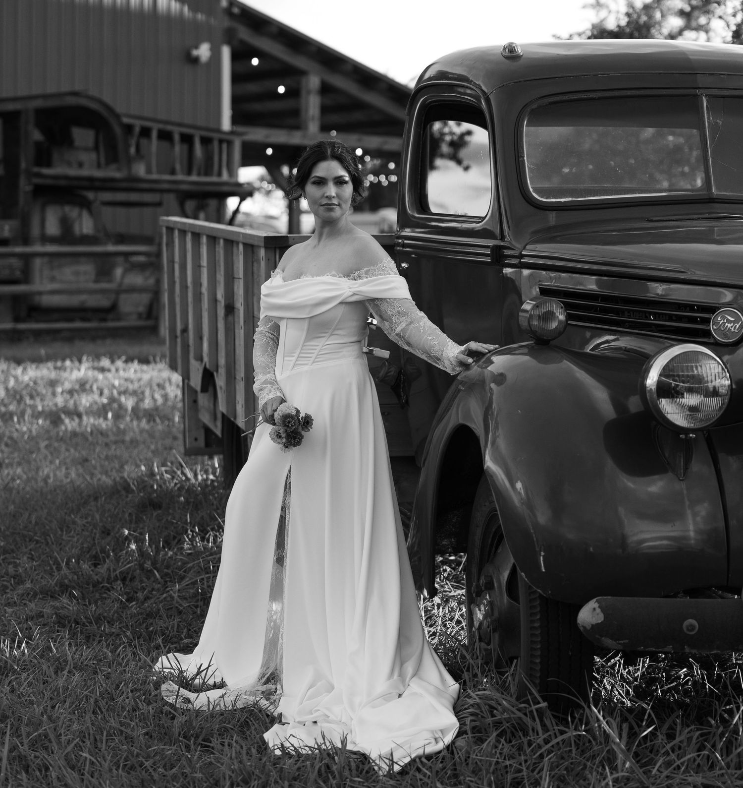 Black and white photo of a bride near vintage cars in a rustic outdoor setting.