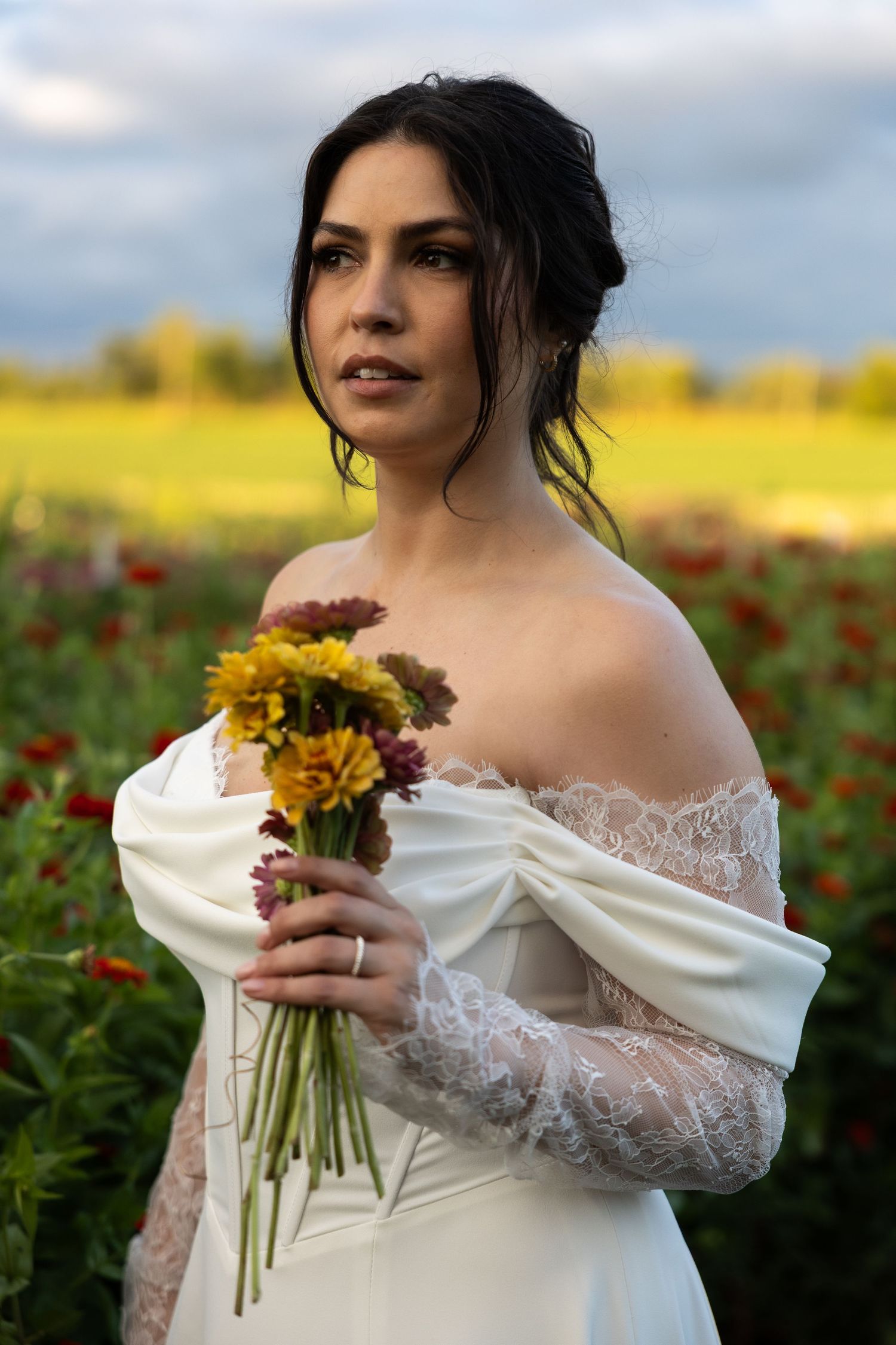 A bride in an off-shoulder lace wedding dress holds a bouquet of sunflowers in a flower field at sunset.