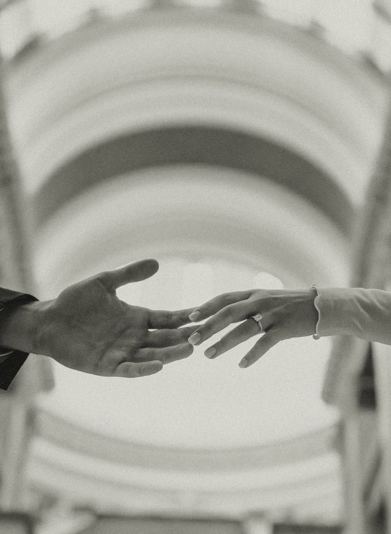 Black and white photo of two hands reaching toward each other beneath an arched ceiling, one wearing a bracelet.