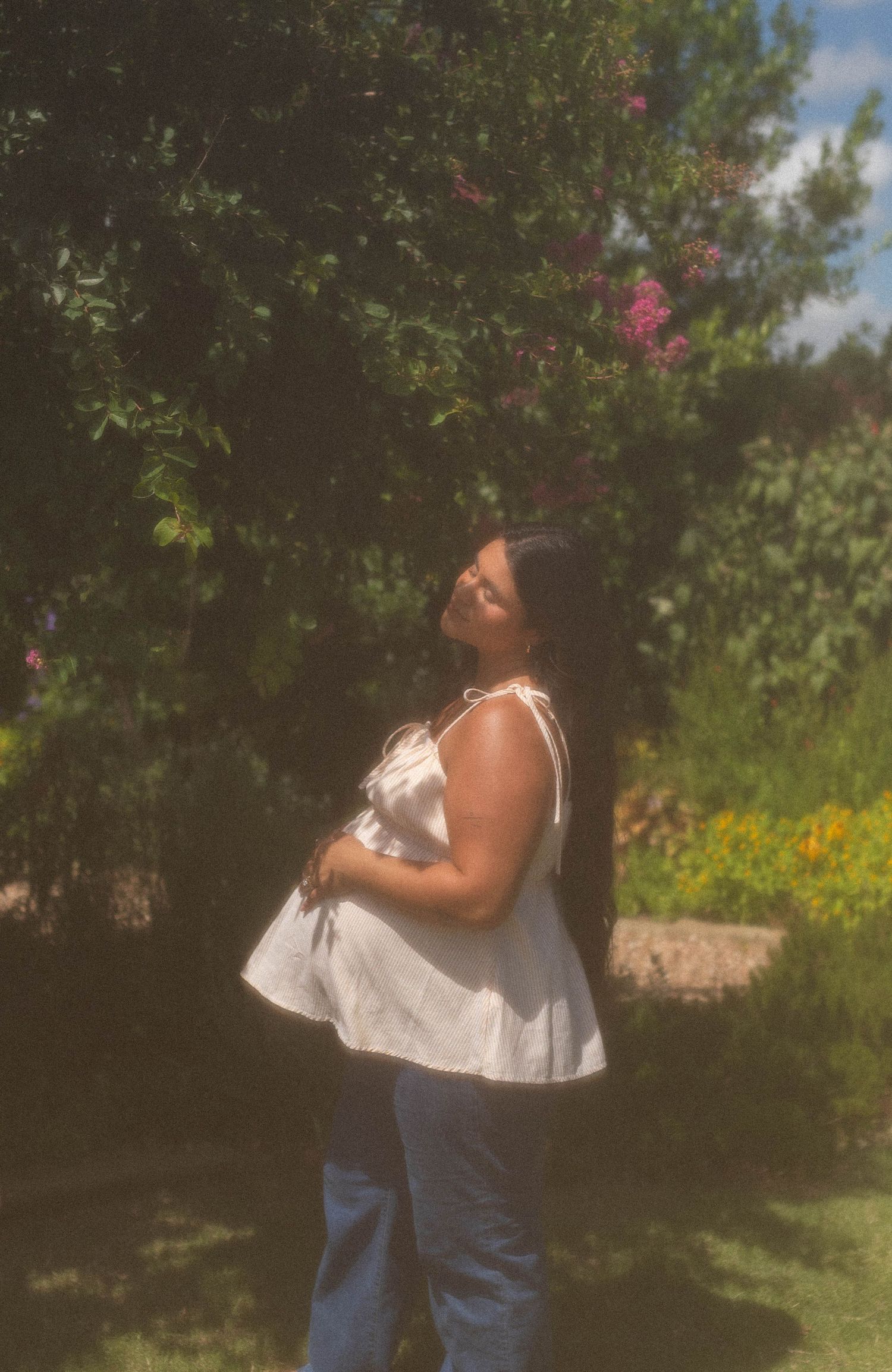 Woman in white top and jeans stands in sunny garden, looking up near blooming trees.