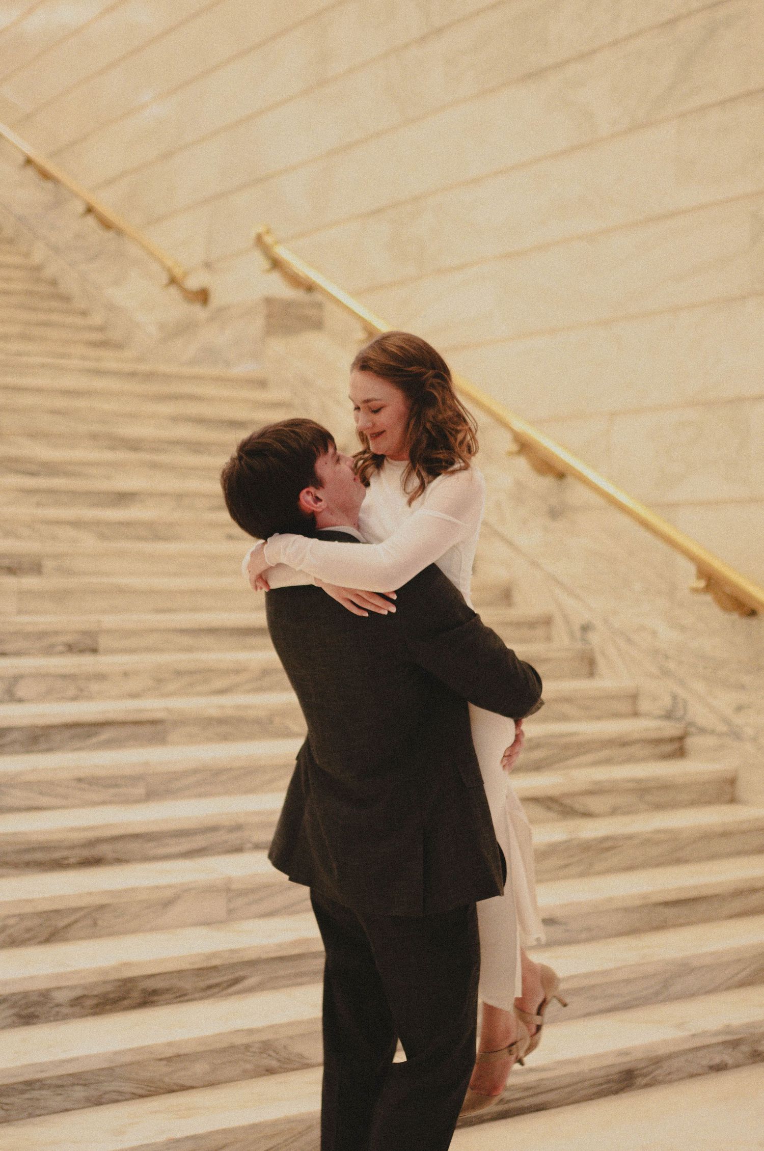 A couple embraces joyfully on marble stairs, the woman in white lifted by her partner in a dark suit.