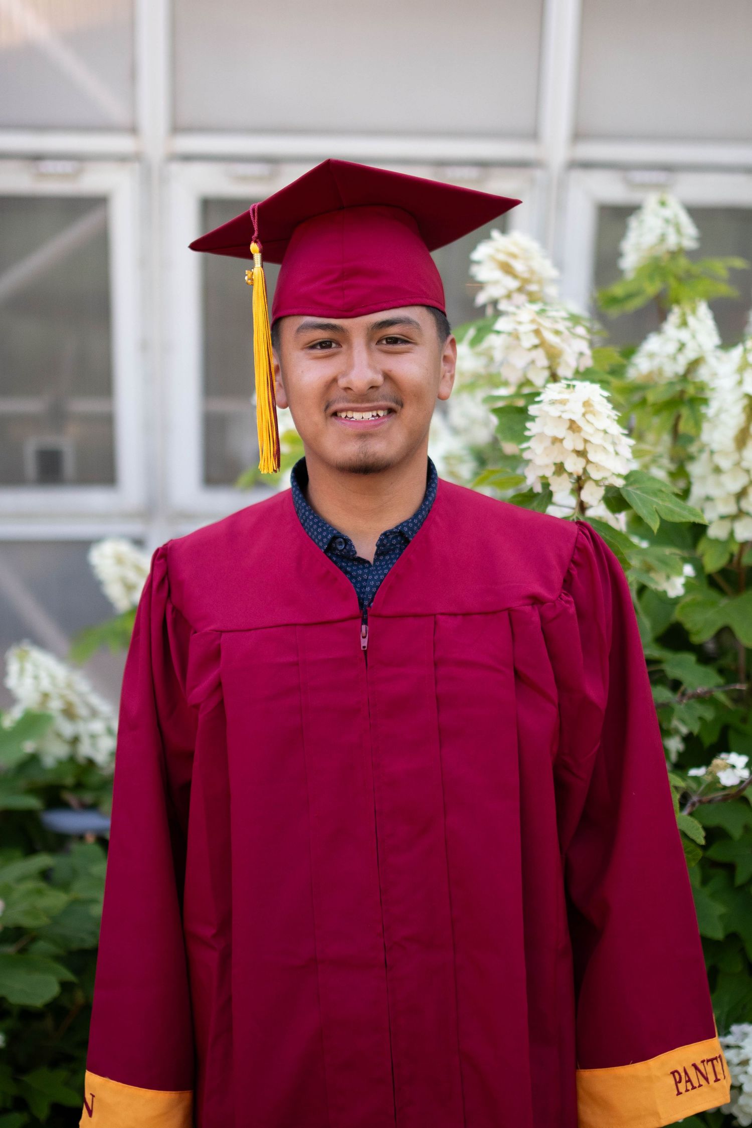 Graduate in burgundy cap and gown with gold tassel standing near white flowers.