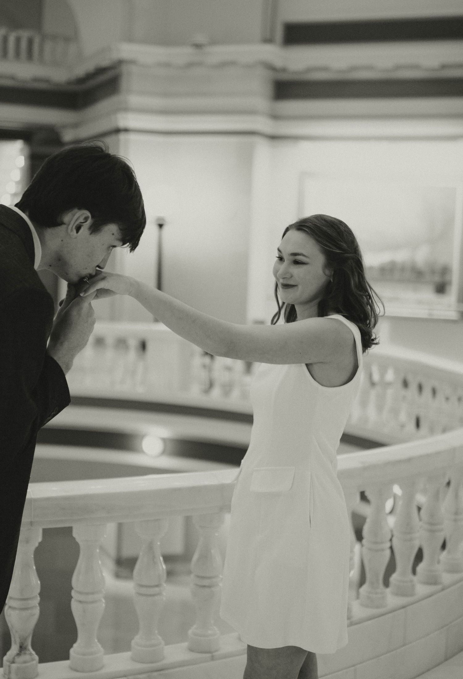 A man kisses a woman's hand on an elegant staircase in a romantic black and white photo.