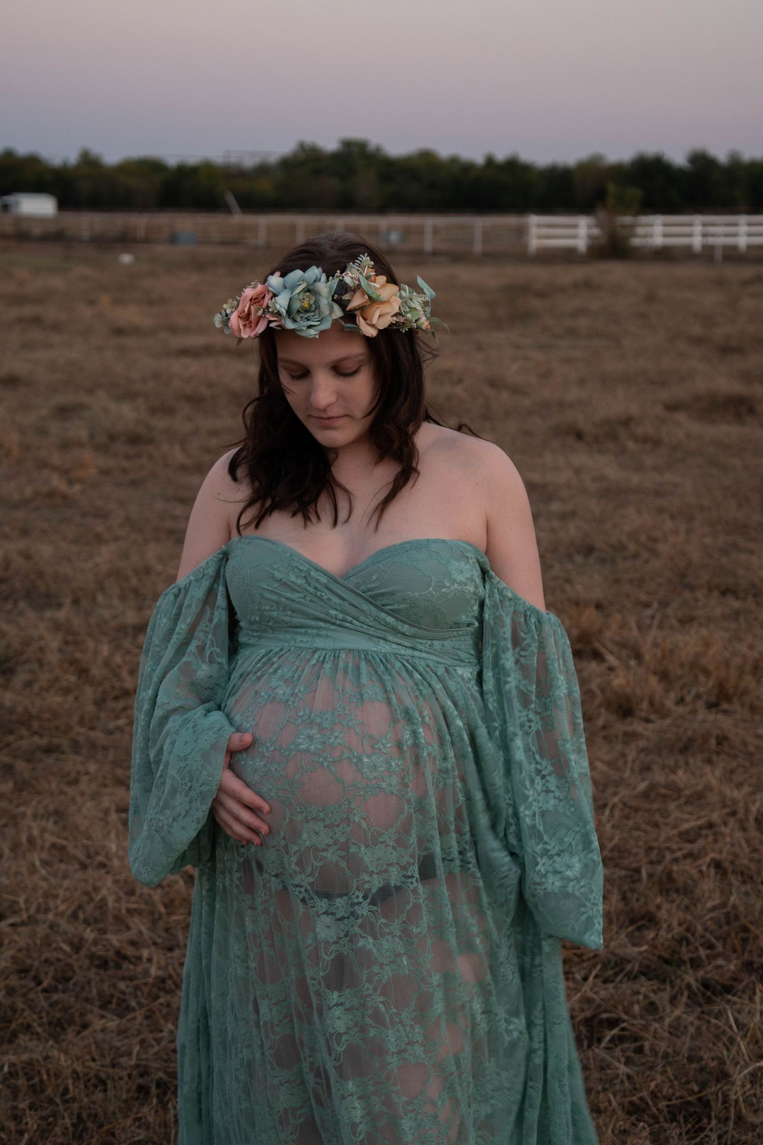Woman in flowing teal lace maternity gown poses in field at sunset.