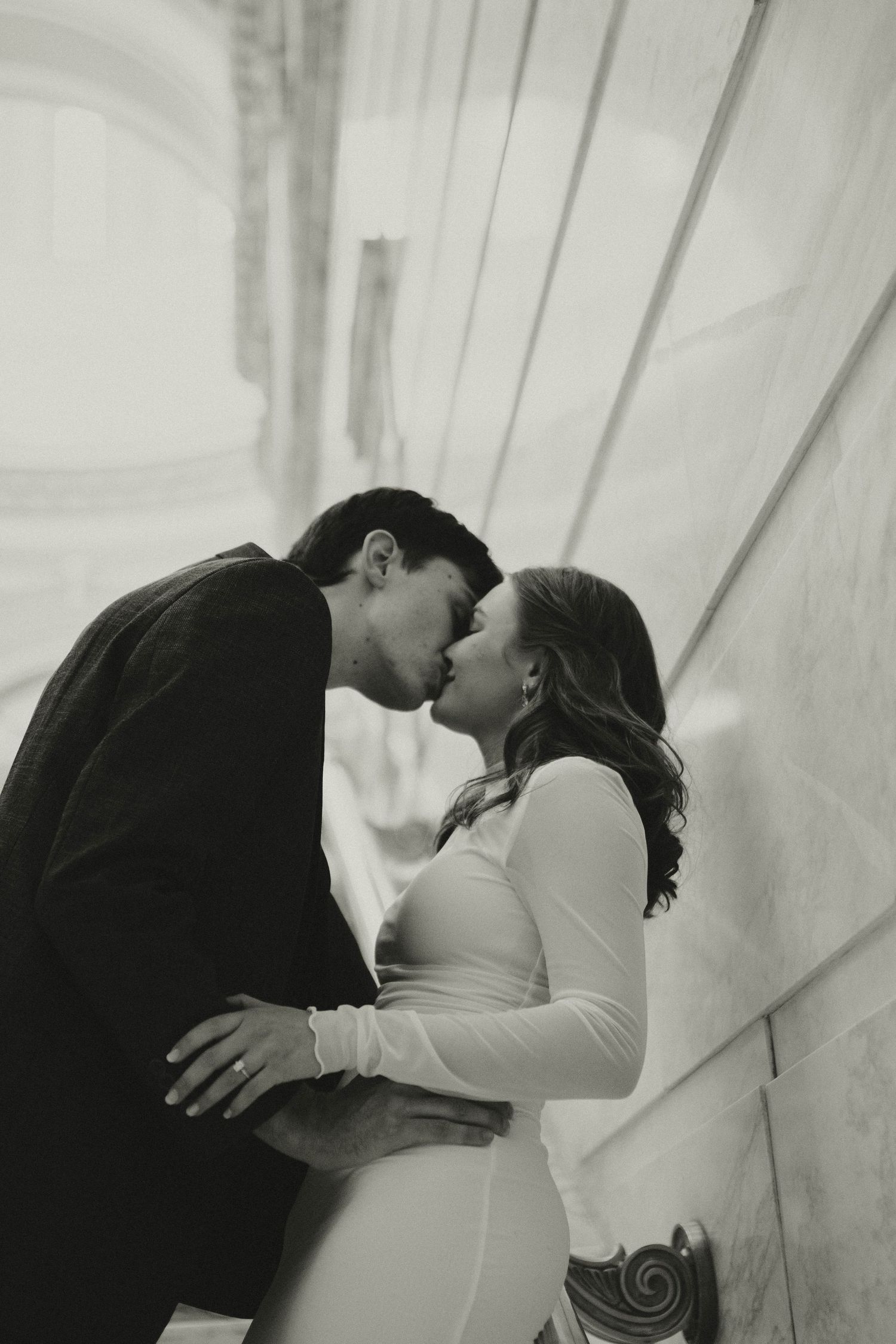 Black and white photo of a couple kissing against a stone wall, woman in white dress and man in dark jacket.