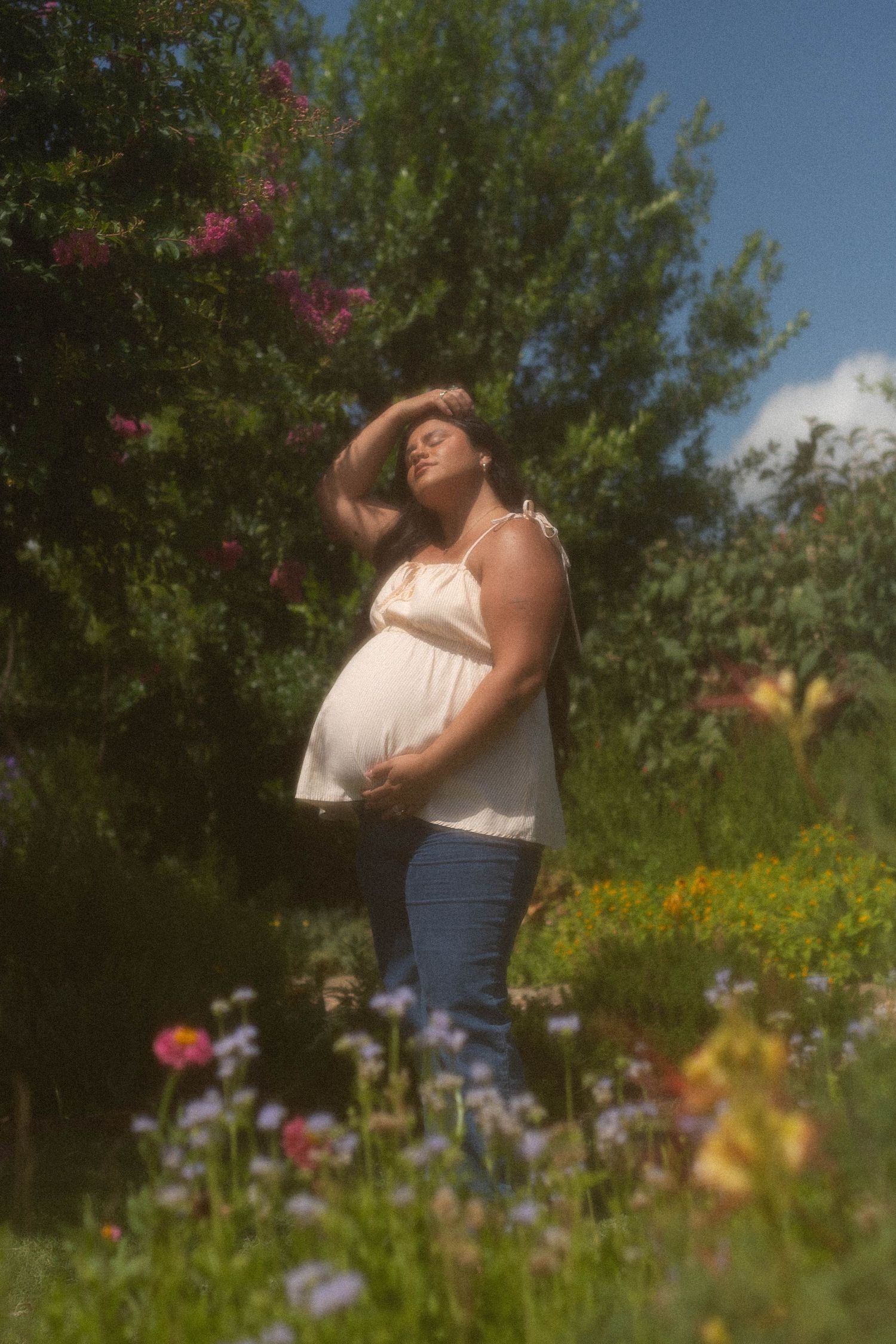 Pregnant woman in white top and jeans cradling her belly among wildflowers in a sunny garden.