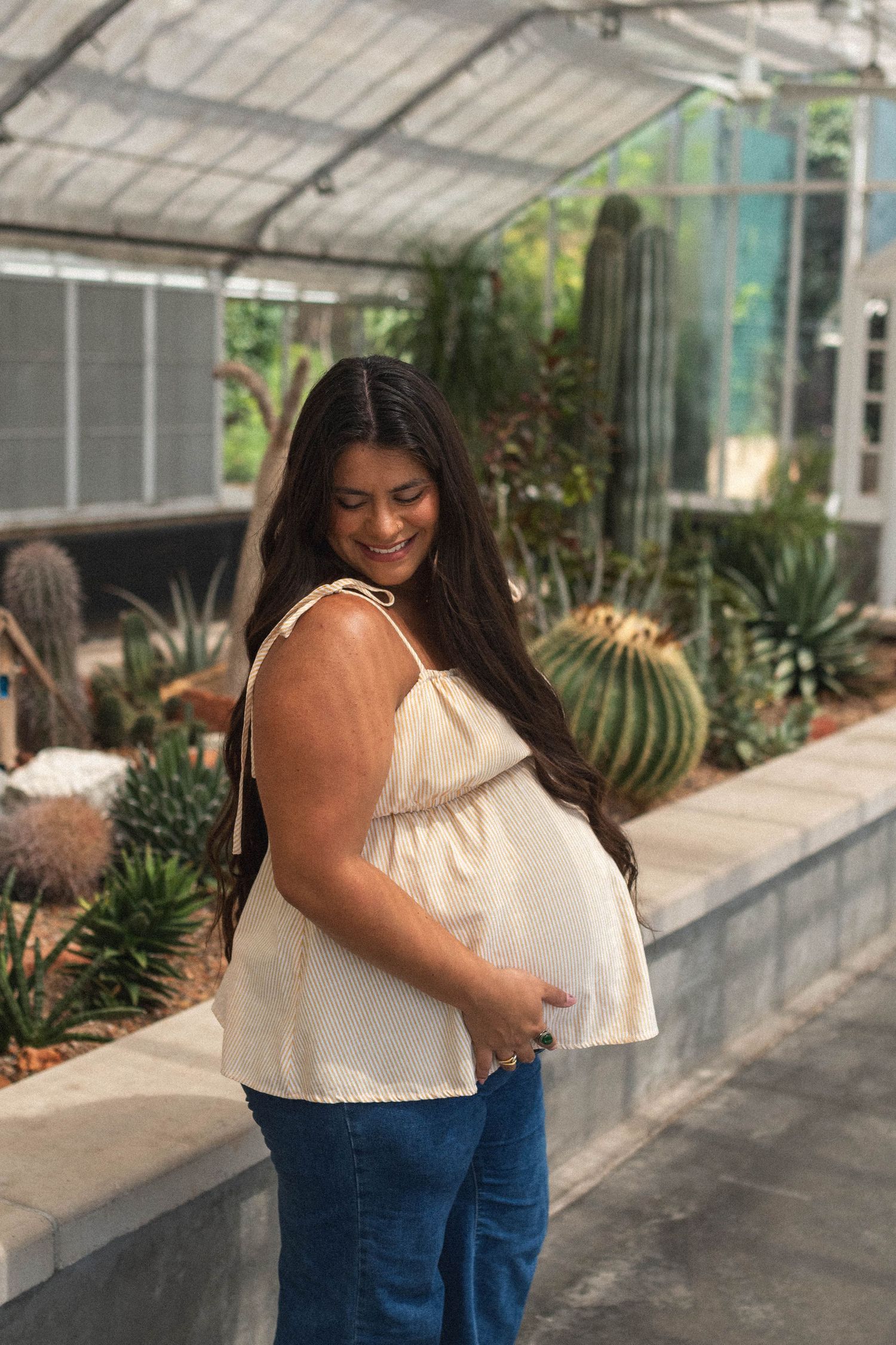 Smiling pregnant woman in beige top and jeans cradling her baby bump in a greenhouse with cacti.
