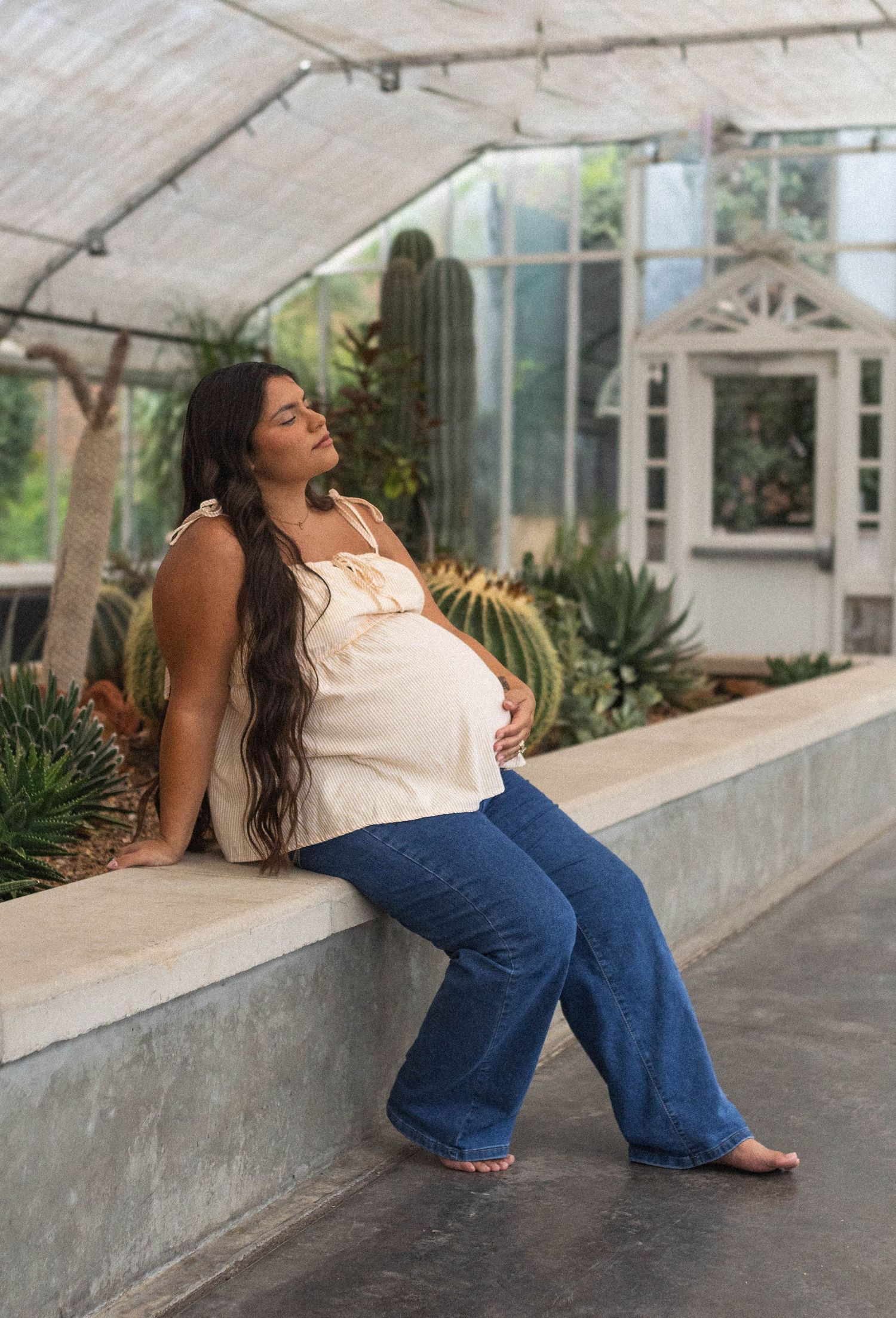 Pregnant woman in white top and flared jeans relaxing in a greenhouse surrounded by cacti and succulents.