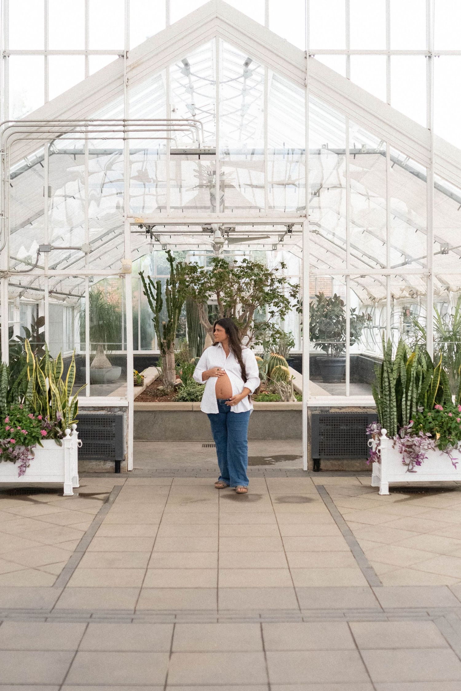 Pregnant woman in white top and blue jeans standing inside a bright greenhouse with lush plants and glass walls.