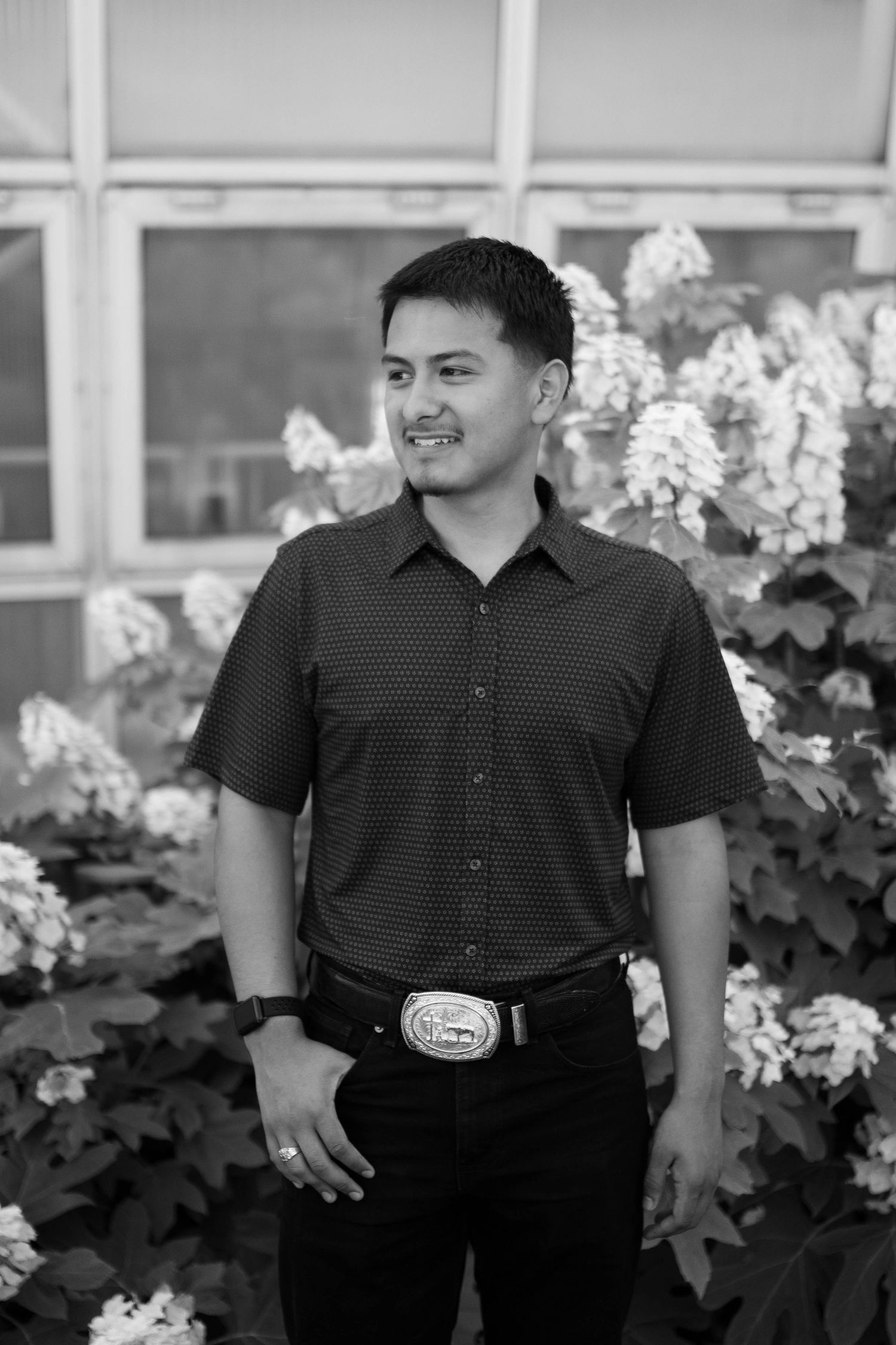 Person in dark polo shirt with decorative belt buckle standing by flowering bushes.
