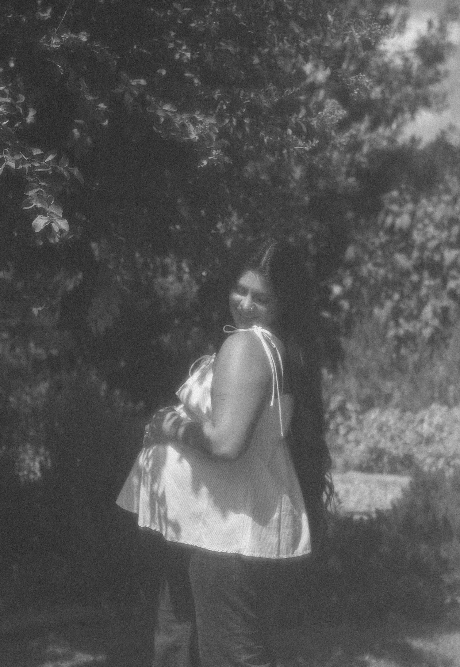 Black and white photo of a pregnant woman in a white top standing outdoors near trees.