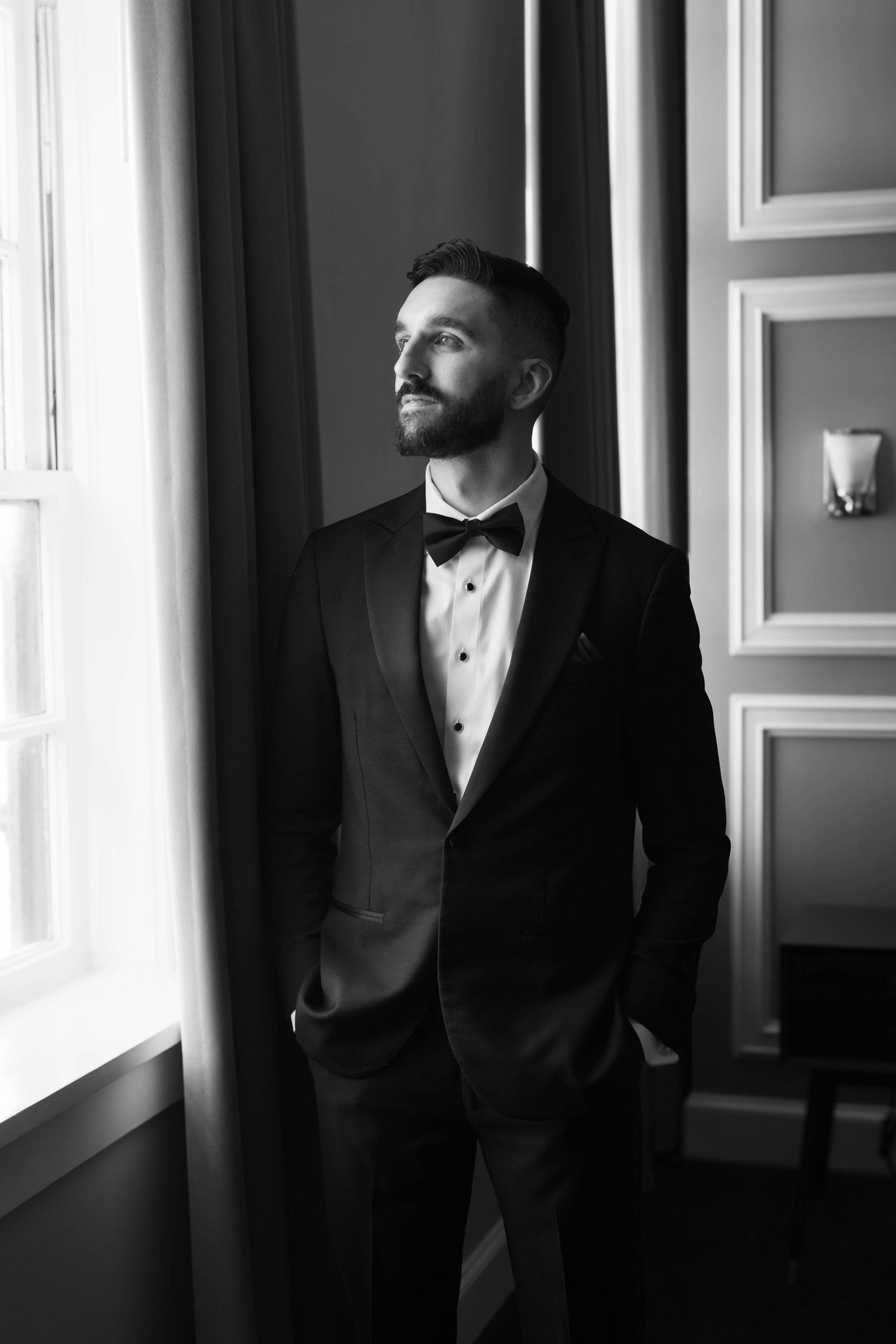 Dramatic black and white portrait of a groom in a tuxedo looking out a window.