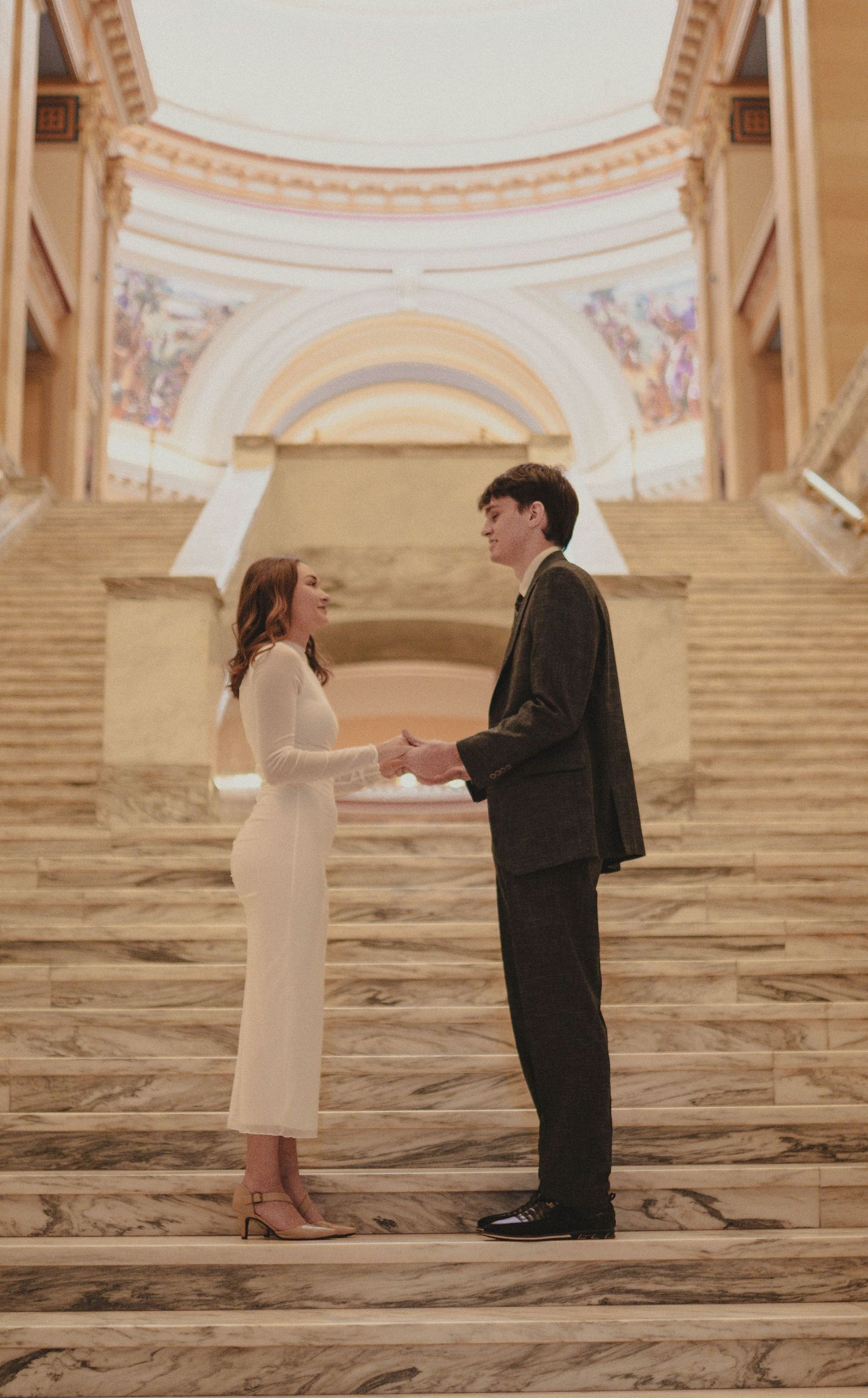 Couple in wedding attire holding hands on marble stairs inside an elegant building with murals.