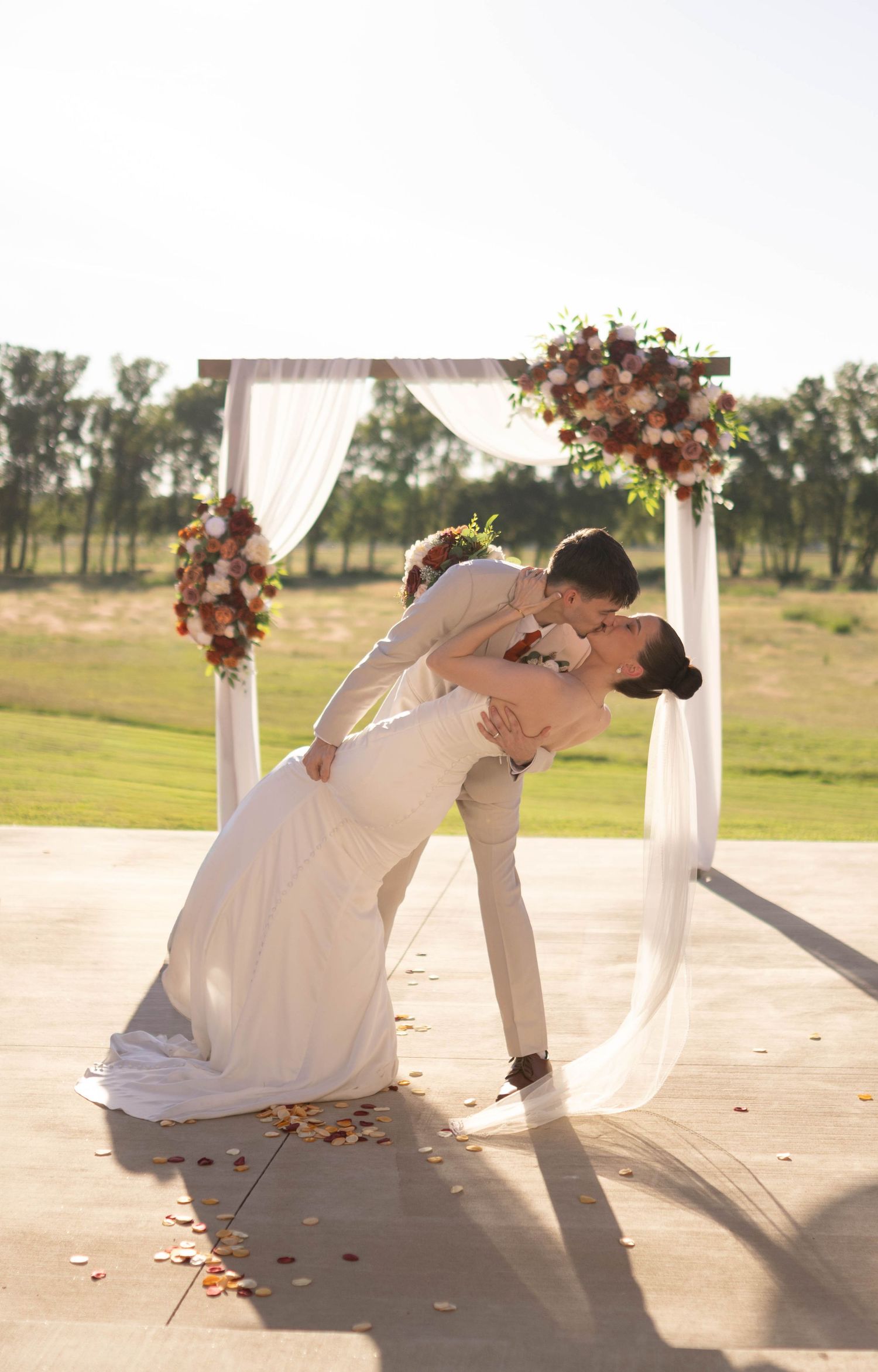 Romantic dip kiss under an elegant wedding arch decorated with burgundy and white flowers.