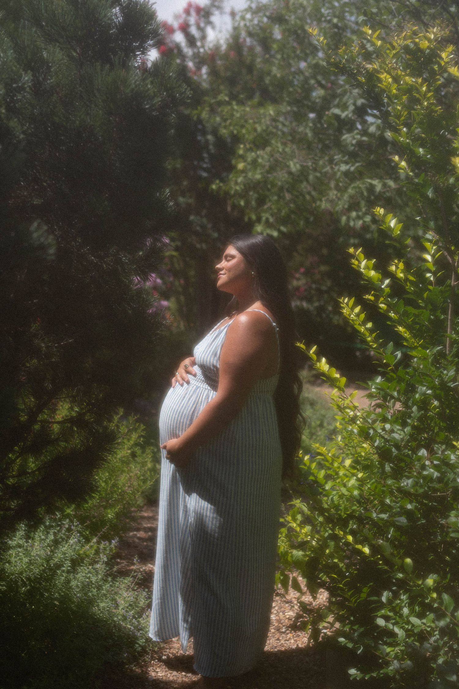 Pregnant woman in striped maxi dress cradling baby bump in lush garden, serene maternity photo in natural sunlight.