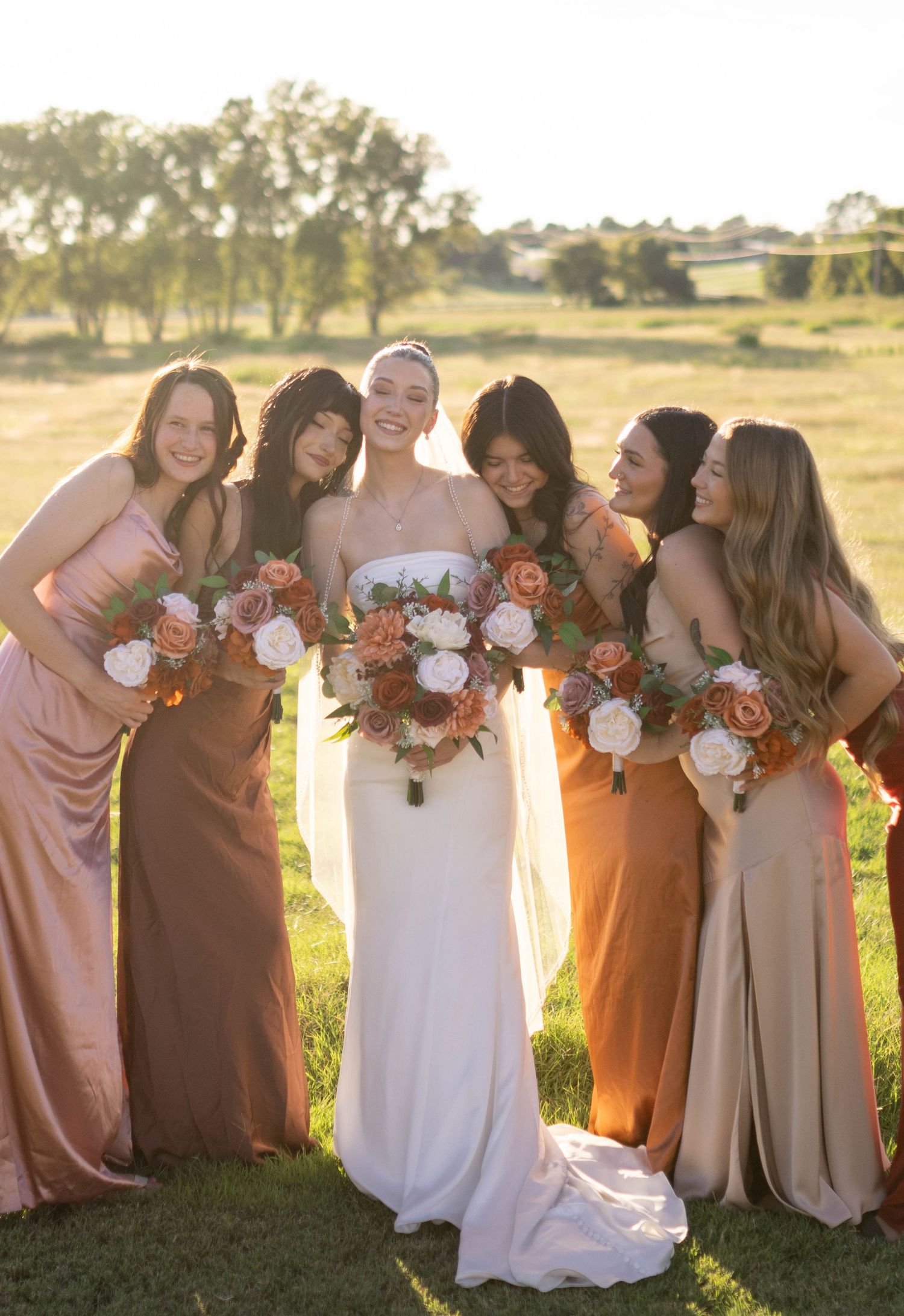 Bridal party in earth-toned dresses poses together in sunset field with bouquets.