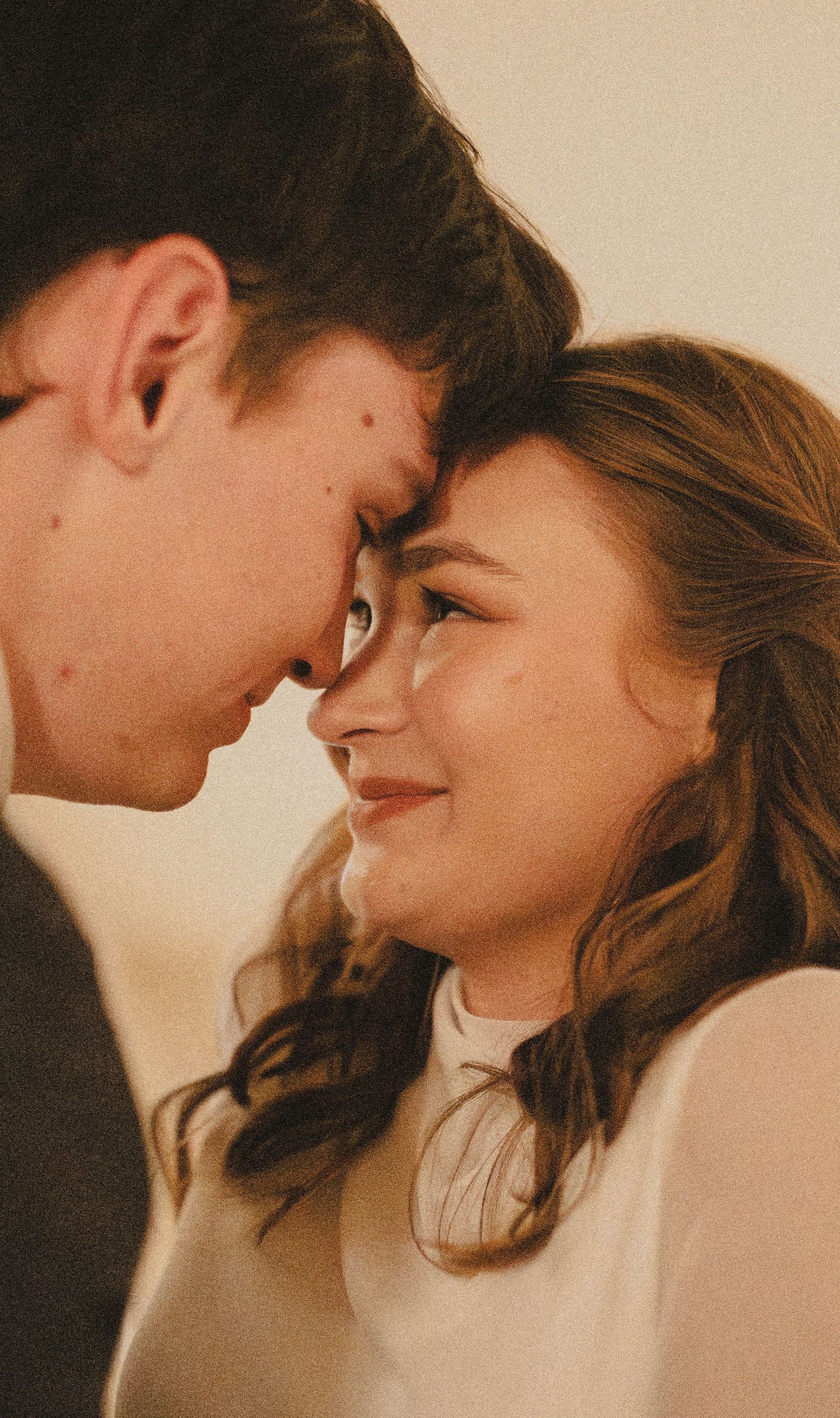 A young couple shares an intimate forehead-to-forehead moment, smiling softly in warm indoor lighting.