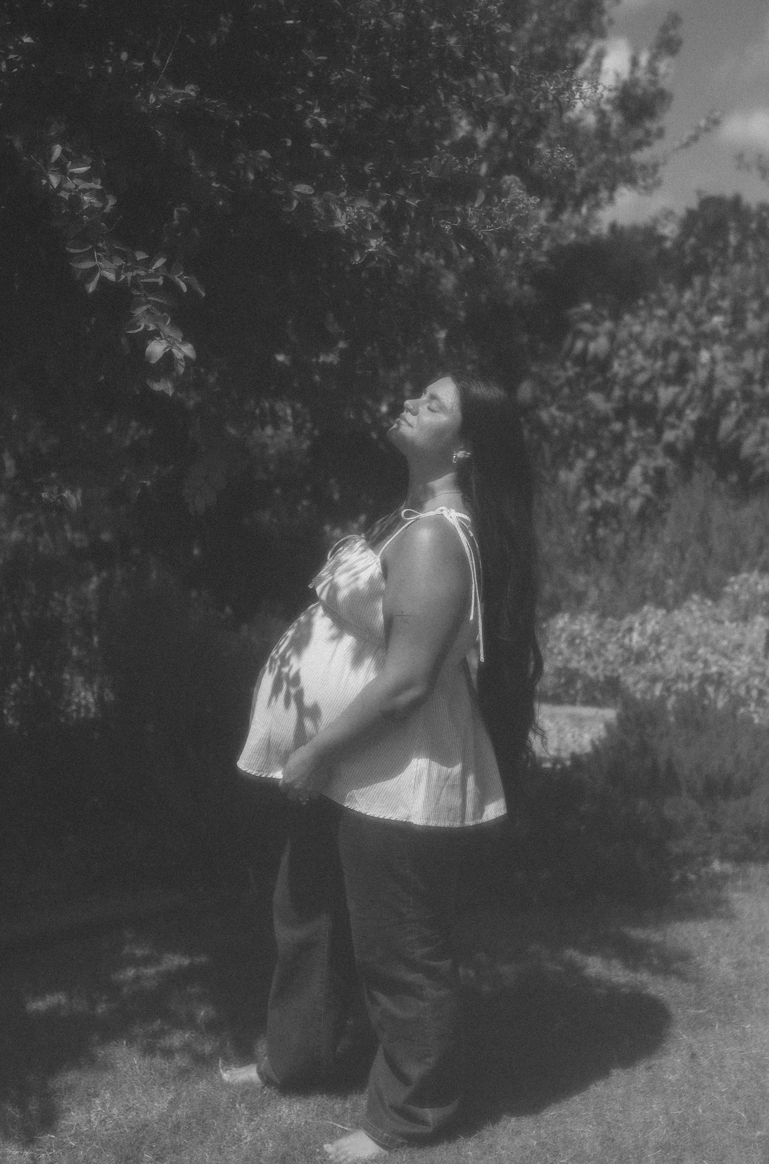 Pregnant woman standing outdoors in black and white photo, looking up with long hair, wearing flowy top and dark pants.