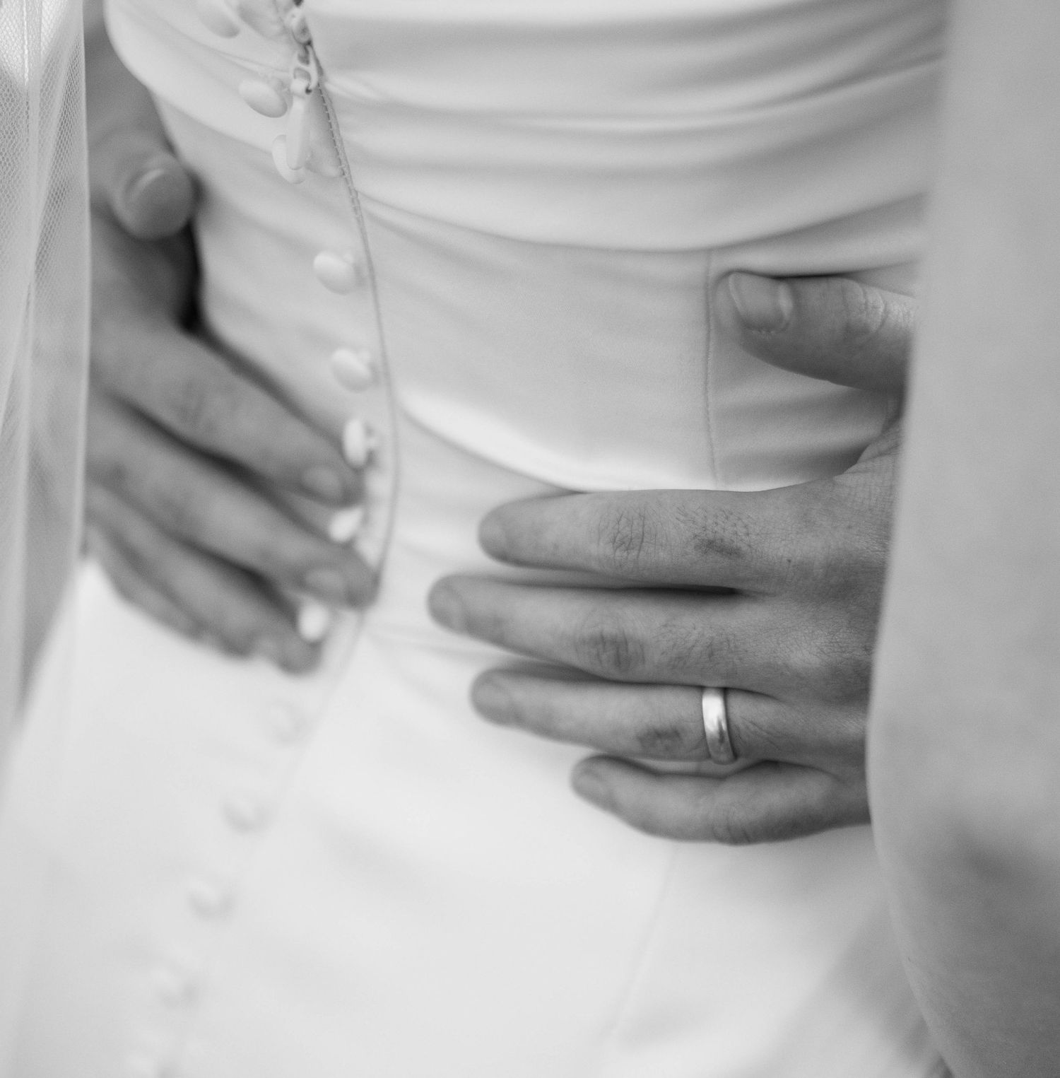 Close-up detail of buttons and hands on a white wedding dress showing wedding ring.