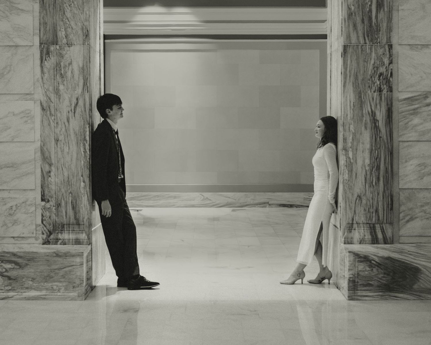 A man and woman in formal attire face each other in a grand marble hallway in a black and white photo.