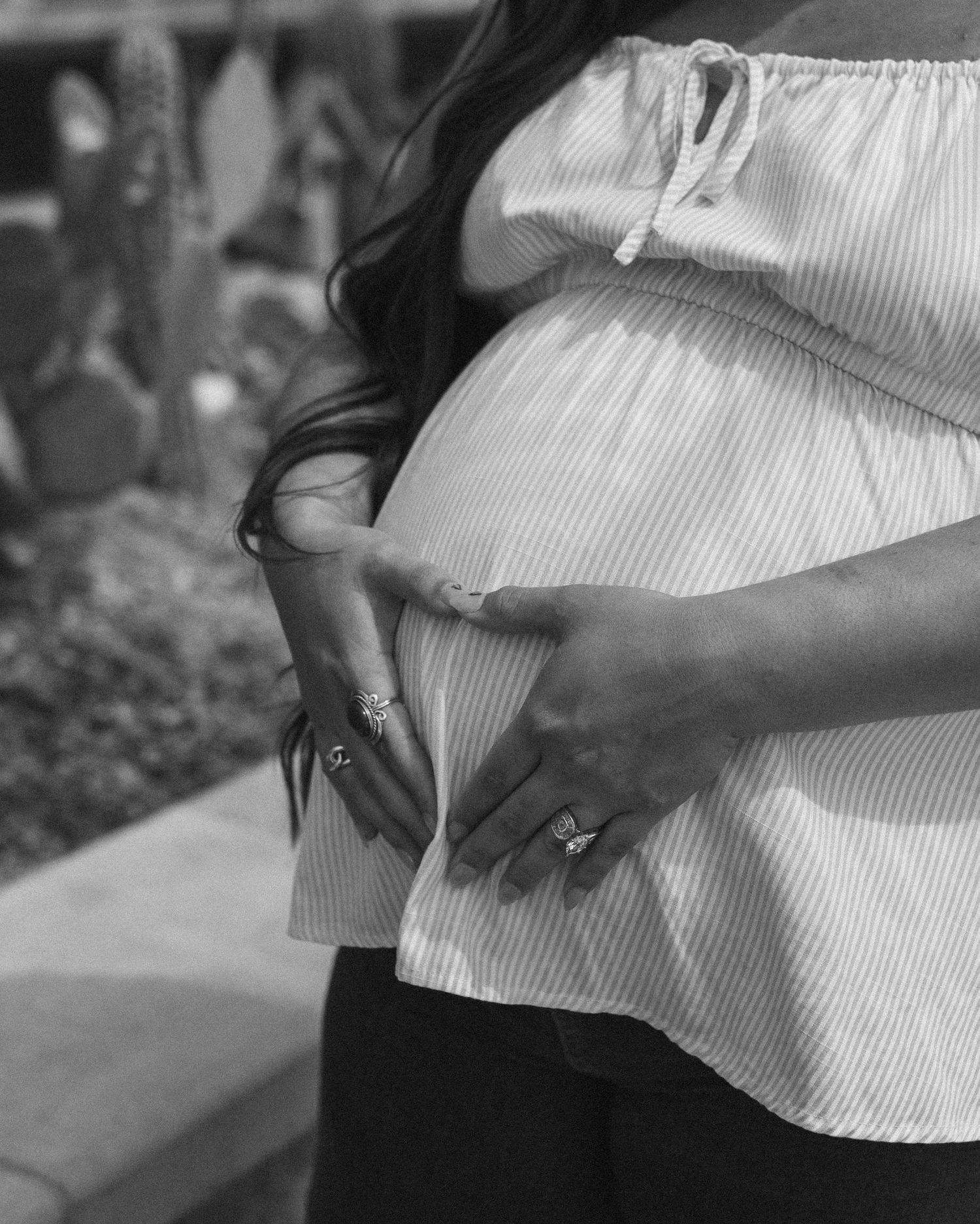Black and white photo of a pregnant woman cradling her baby bump, wearing rings and an off-shoulder striped top.