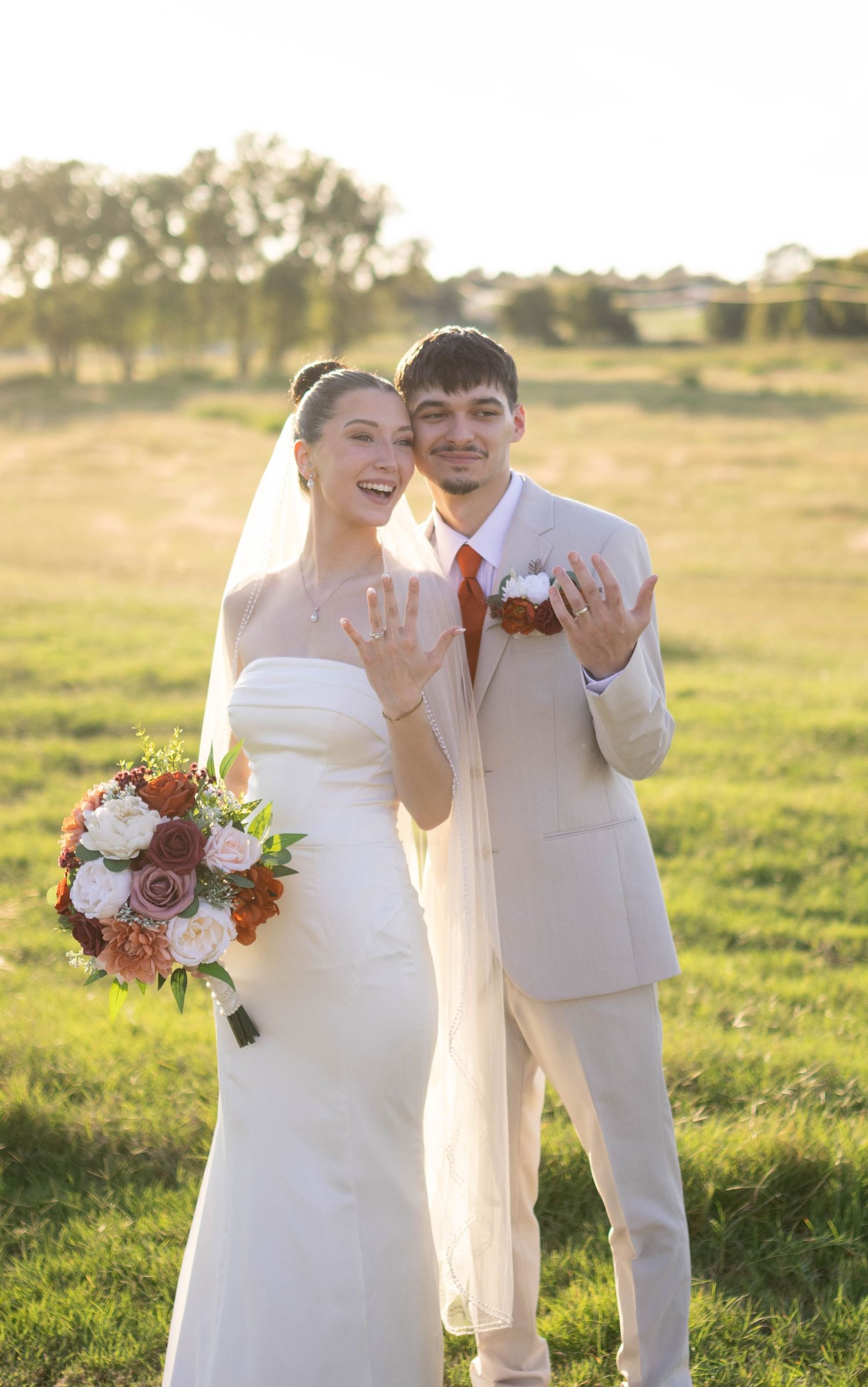 Bride in white dress and groom in light suit pose in golden sunlit field with bouquet.