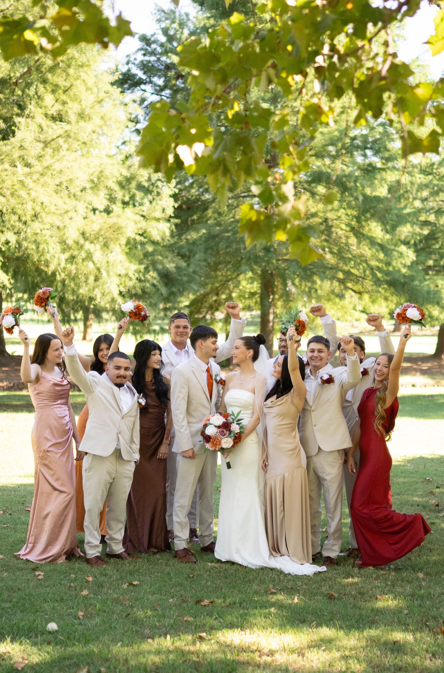 Large wedding party in blush, burgundy and beige poses together under trees for a fun group photo.